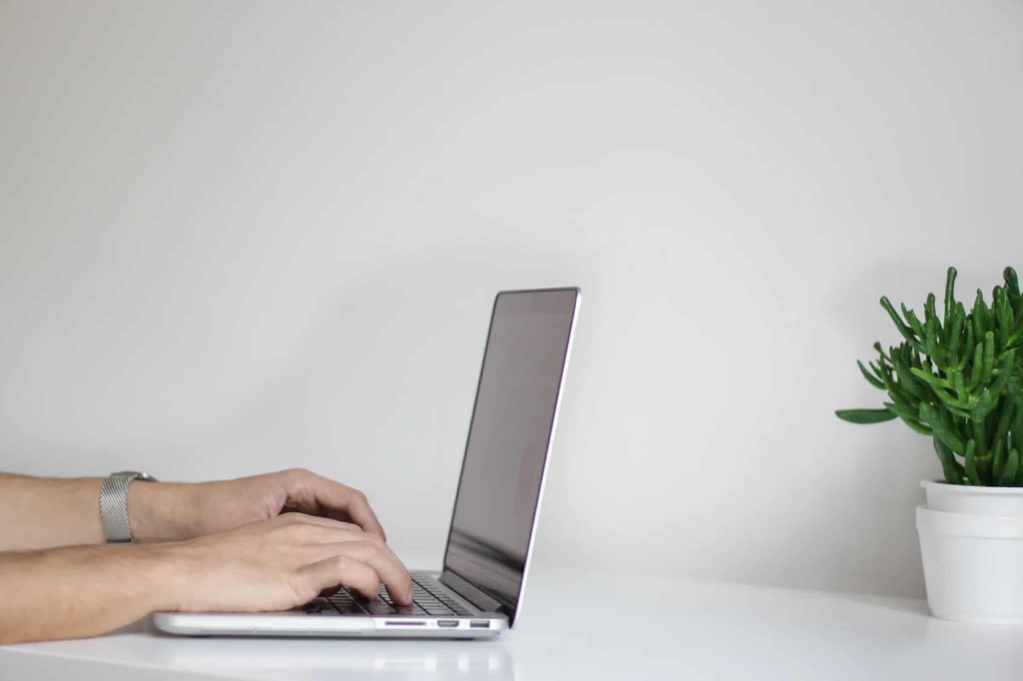 Close-up of a person's hands typing on a laptop keyboard at a clean white desk with a small potted succulent nearby.
