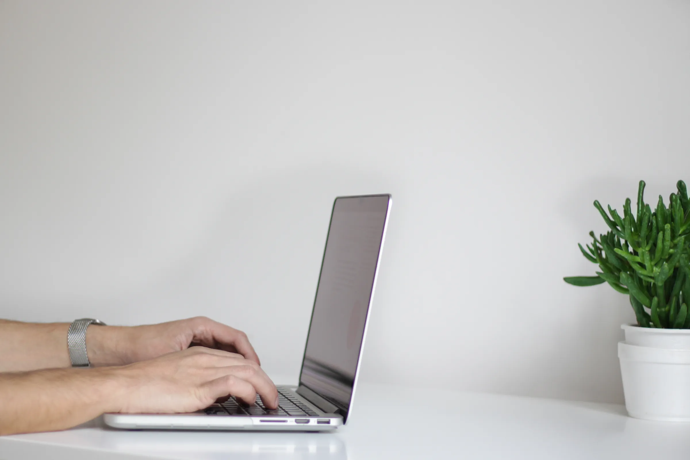 Close-up of a person's hands typing on a laptop keyboard at a clean white desk with a small potted succulent nearby.