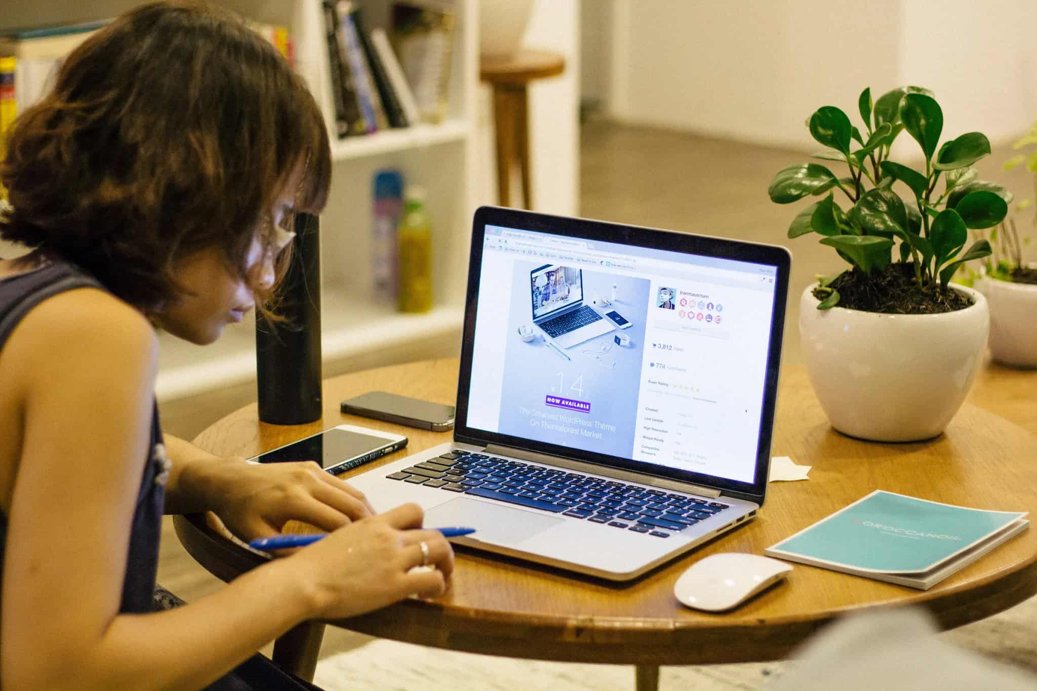 A person sits at a small table taking notes by hand while working on a laptop, with a smartphone and houseplant nearby.