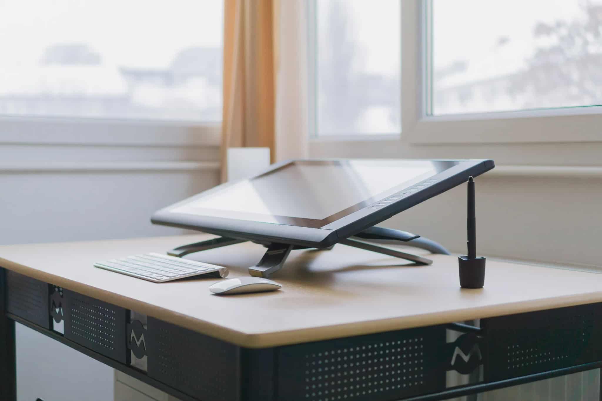 A tilted graphics tablet monitor with stylus sits on a bright, tidy desk beside a wireless keyboard and mouse.