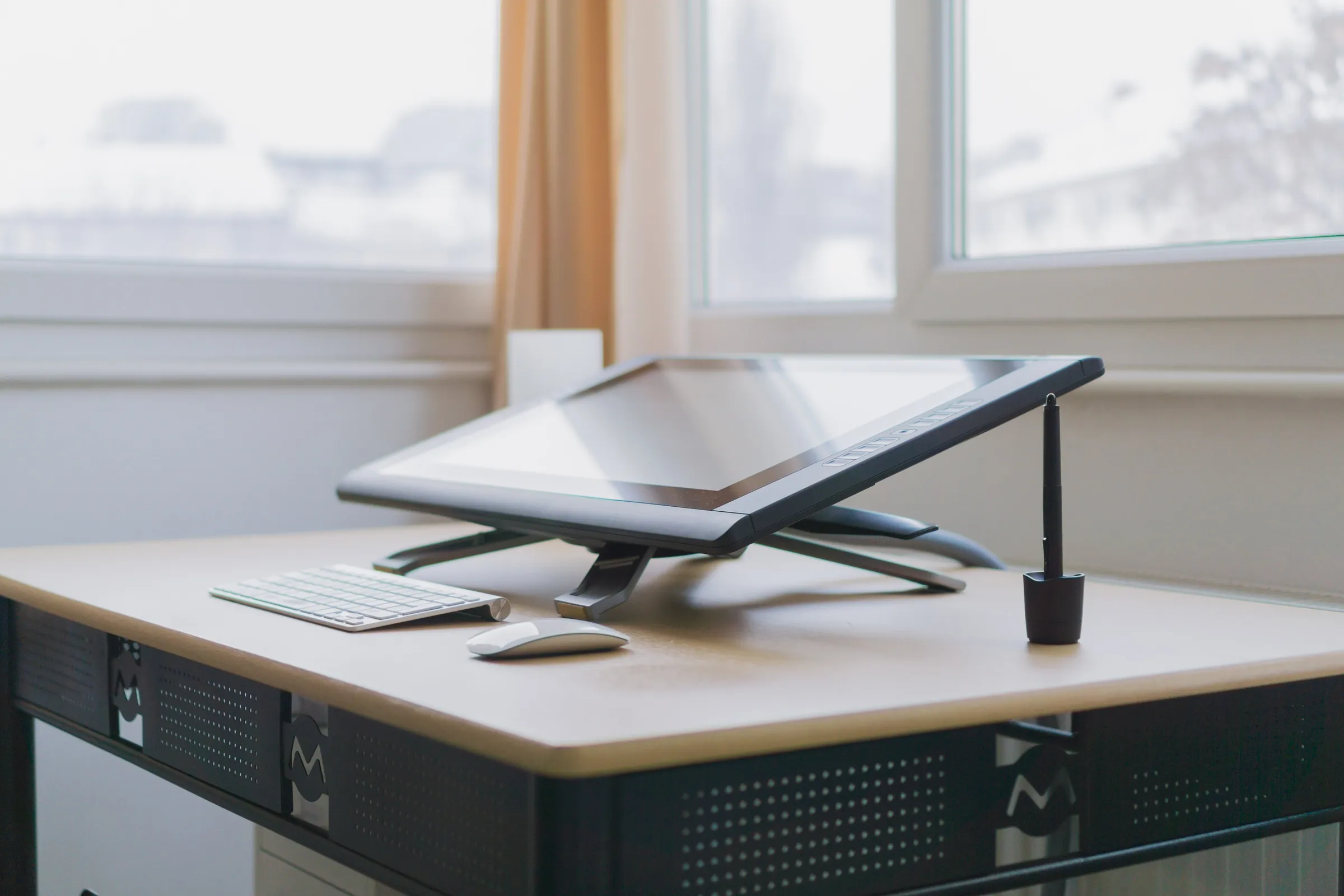A tilted graphics tablet monitor with stylus sits on a bright, tidy desk beside a wireless keyboard and mouse.
