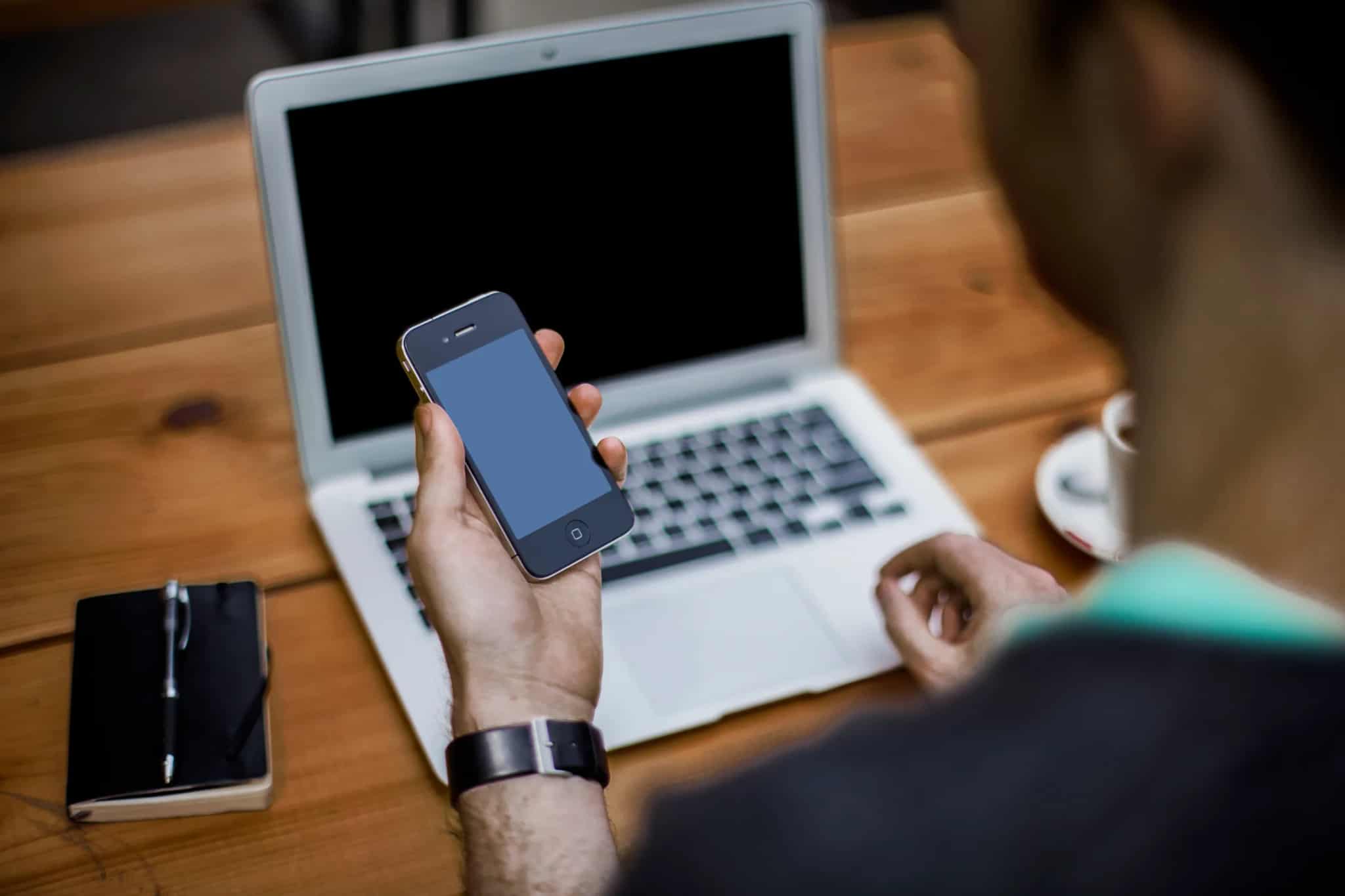 An anonymous person holds a smartphone in one hand while resting the other near an open laptop keyboard on a wooden desk.