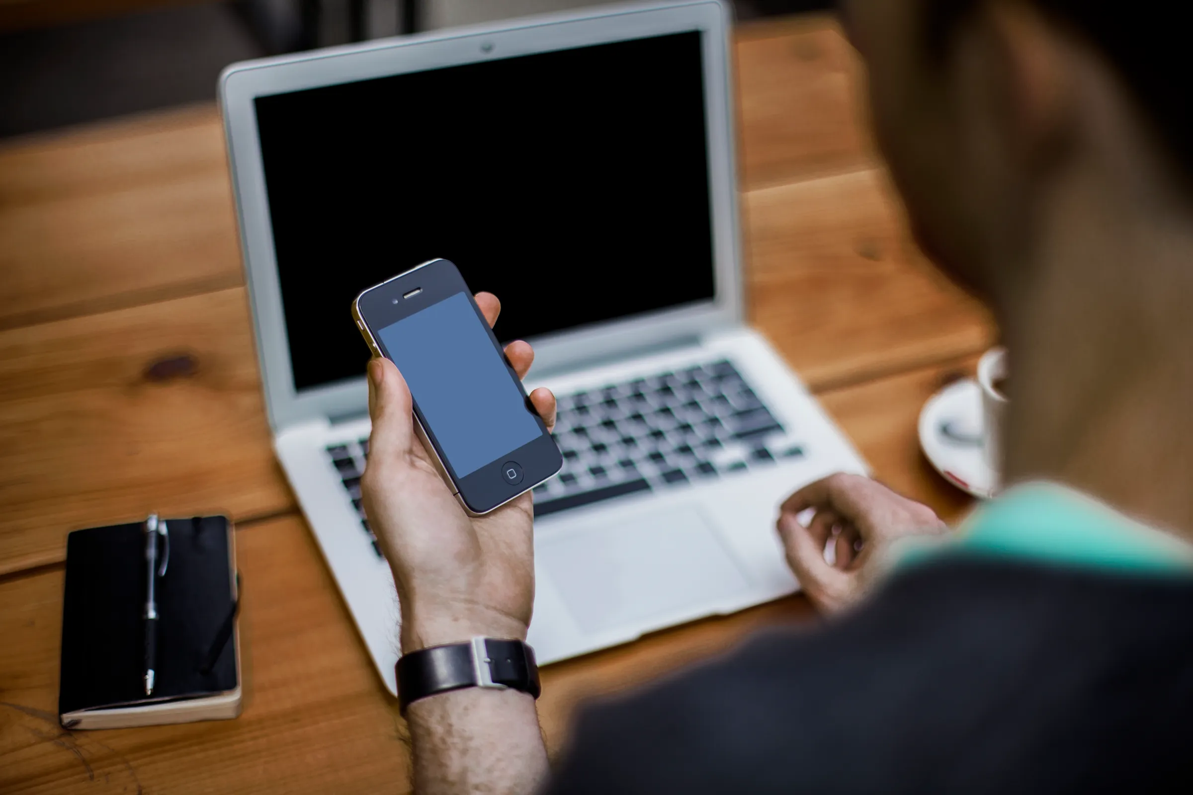An anonymous person holds a smartphone in one hand while resting the other near an open laptop keyboard on a wooden desk.