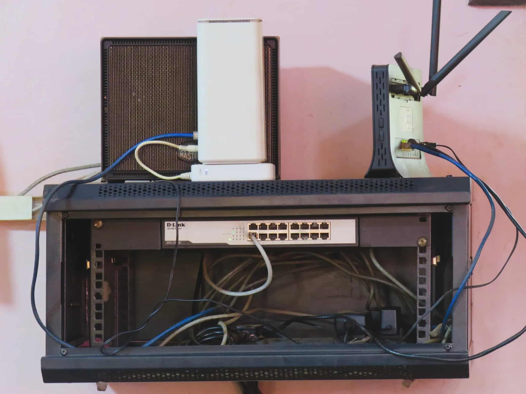 A wall-mounted network rack holding a router, a modem and a 16-port switch with a tangle of ethernet cables running to it.