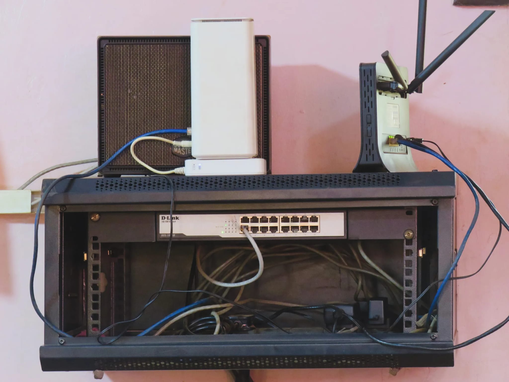 A wall-mounted network rack holding a router, a modem and a 16-port switch with a tangle of ethernet cables running to it.