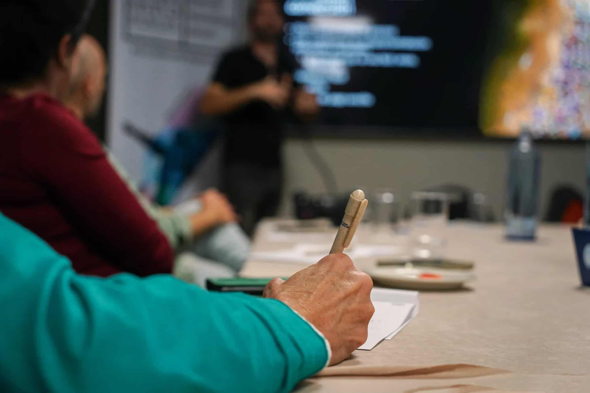 A colleague takes notes at a conference table while a presenter gestures near a large screen during a business meeting.