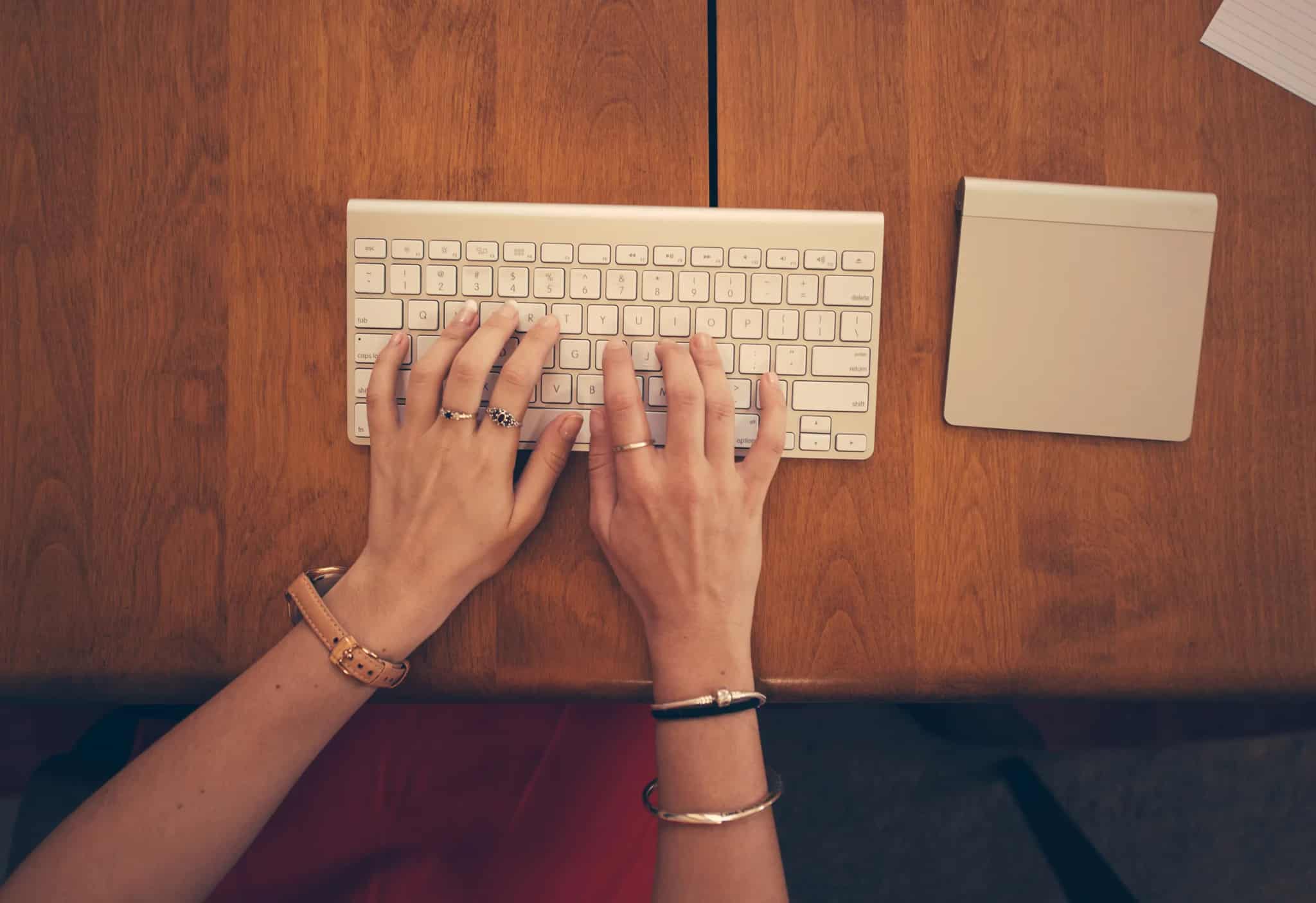 Close-up overhead view of a person's hands typing on a wireless keyboard at a wooden desk, with a piece of paper visible nearby.