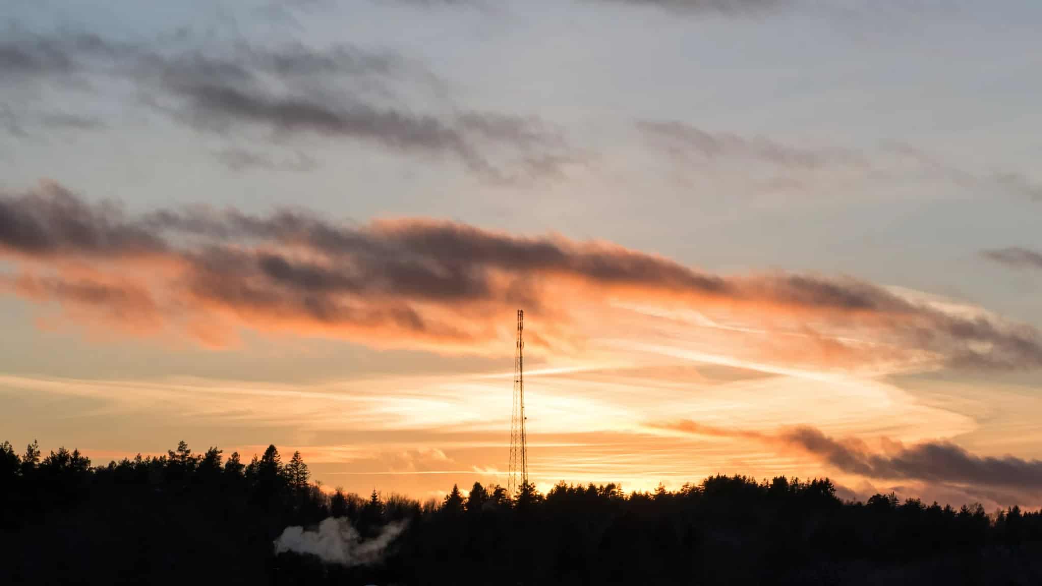 A telecommunications transmission mast silhouetted against a colorful sunset sky above a treeline.