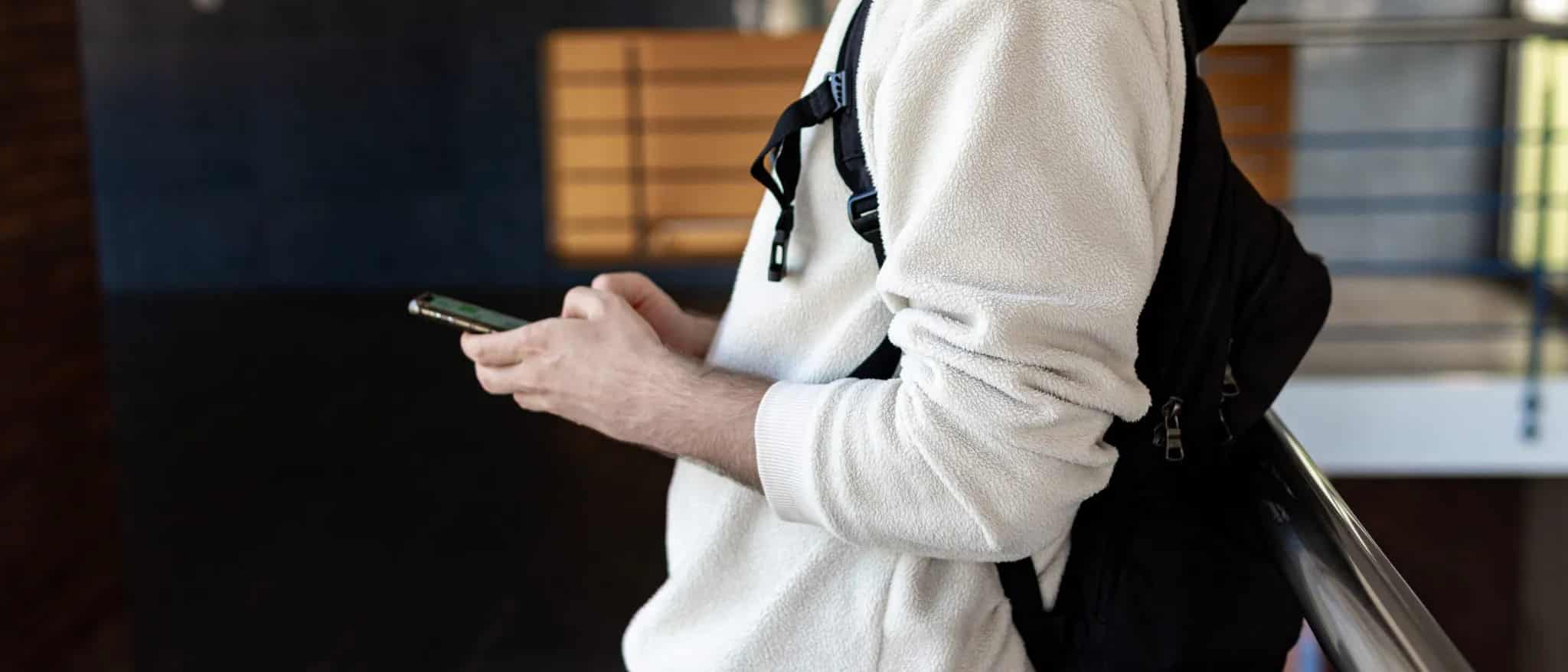 A man in a white backpack and fleece jacket leans against a railing while looking at a smartphone in his hand.