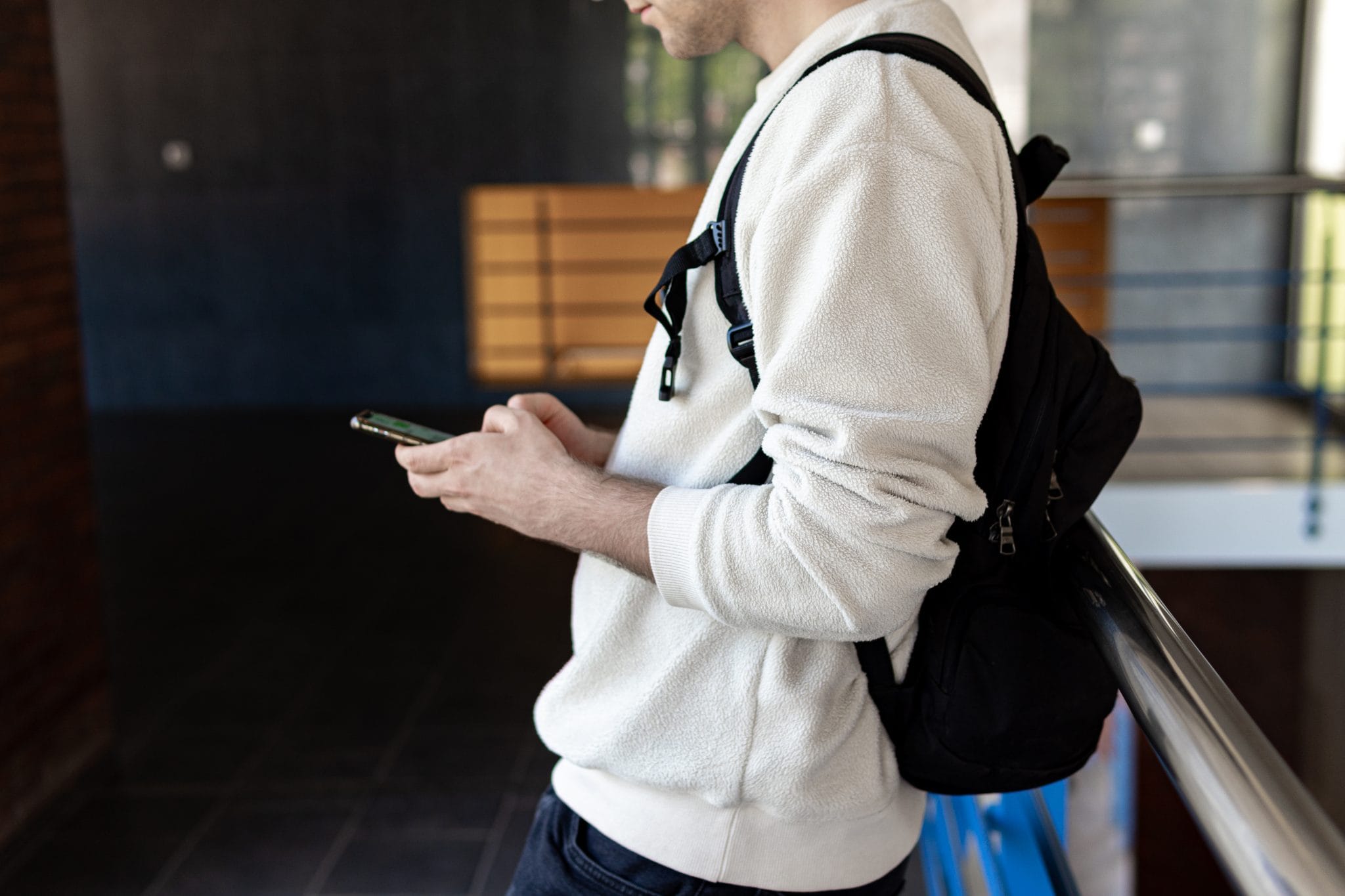 A man in a white backpack and fleece jacket leans against a railing while looking at a smartphone in his hand.