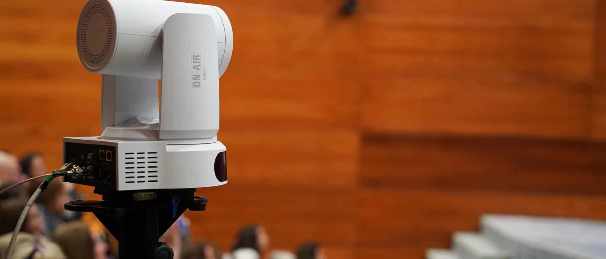 A white broadcast video camera on a tripod pointed at an audience seated in a wood-paneled auditorium.