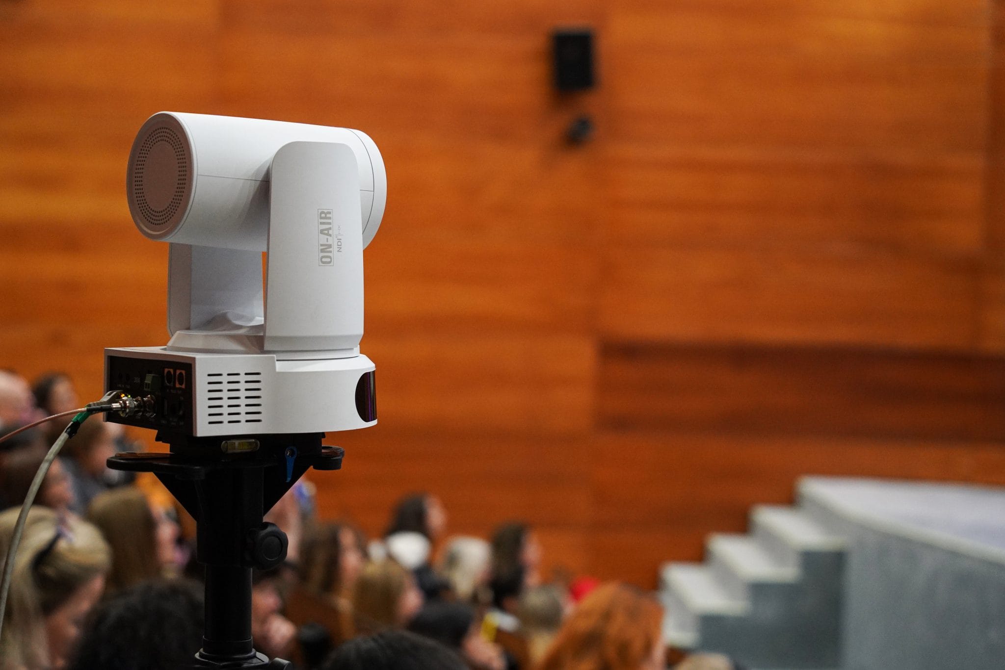 A white broadcast video camera on a tripod pointed at an audience seated in a wood-paneled auditorium.
