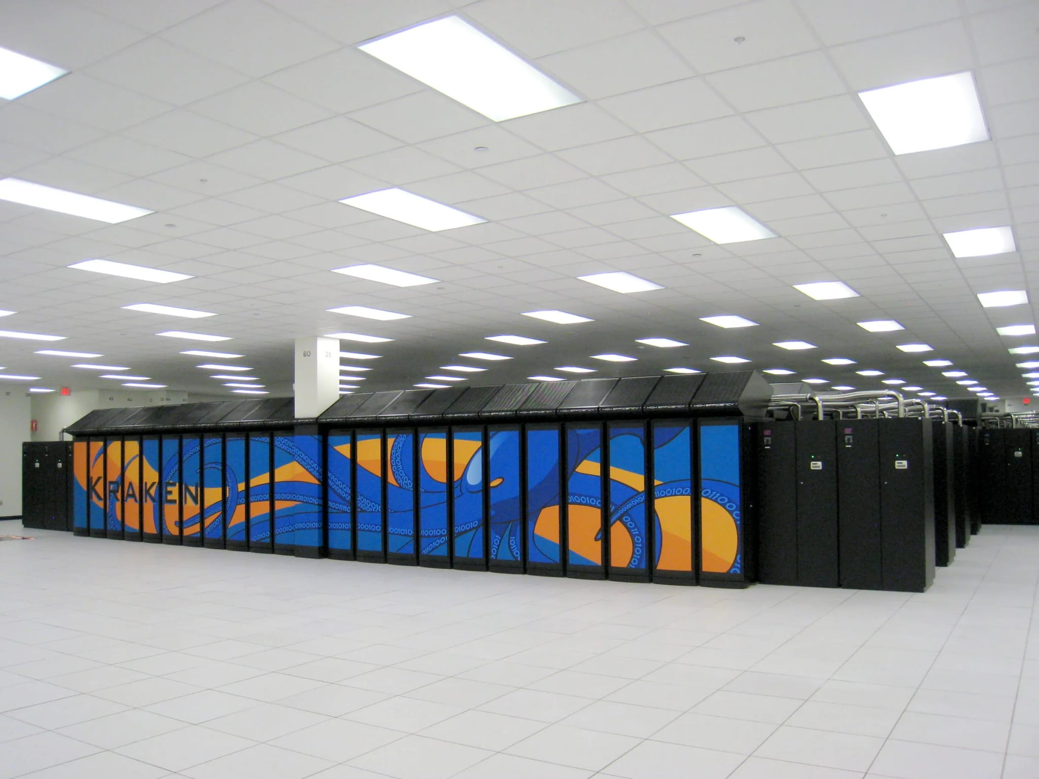 A long row of tall black server cabinets stretching down an aisle of a large data center under fluorescent lighting.