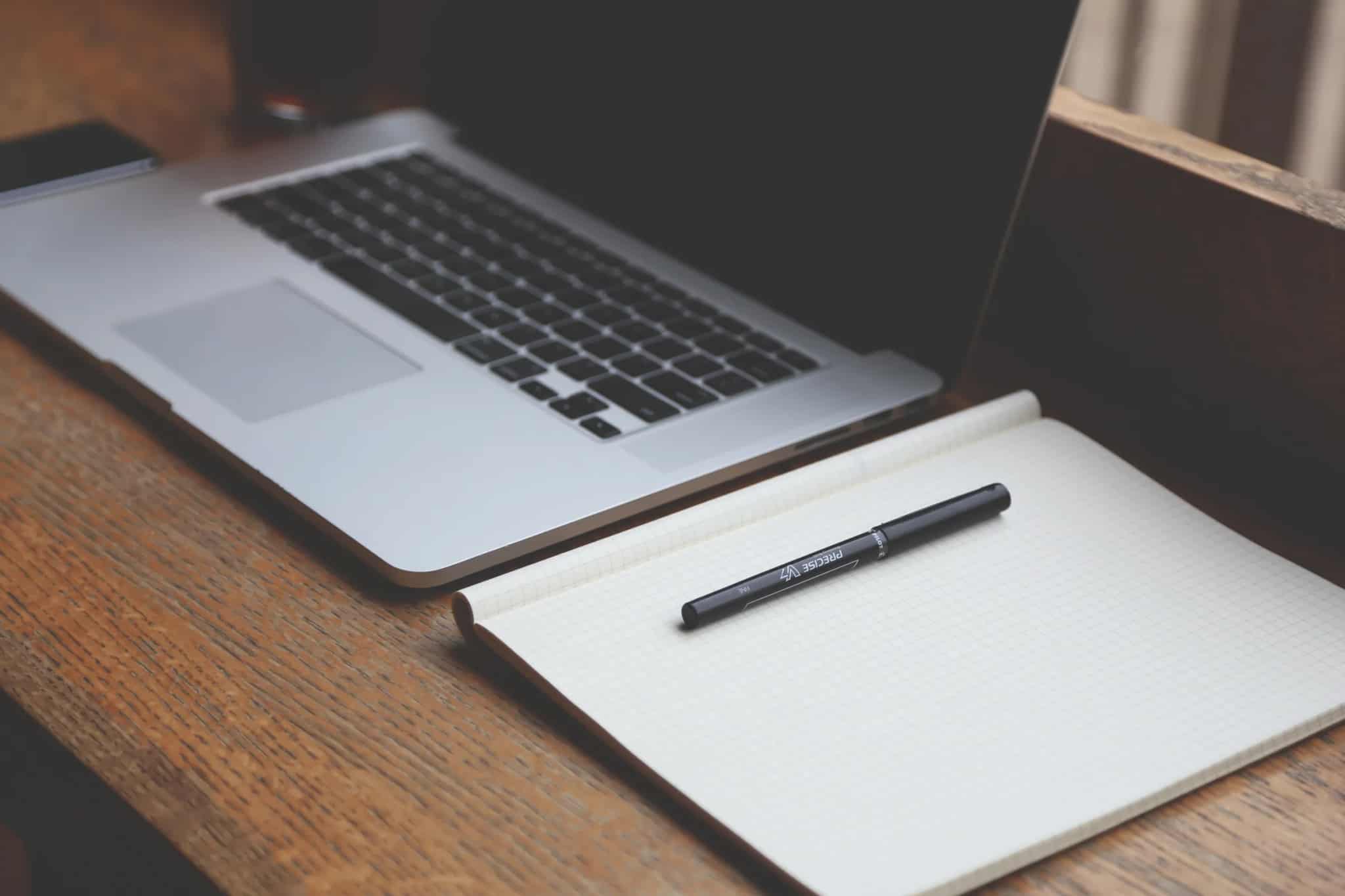 A laptop and an open blank notebook with a pen resting on it, sitting on a wooden desk.