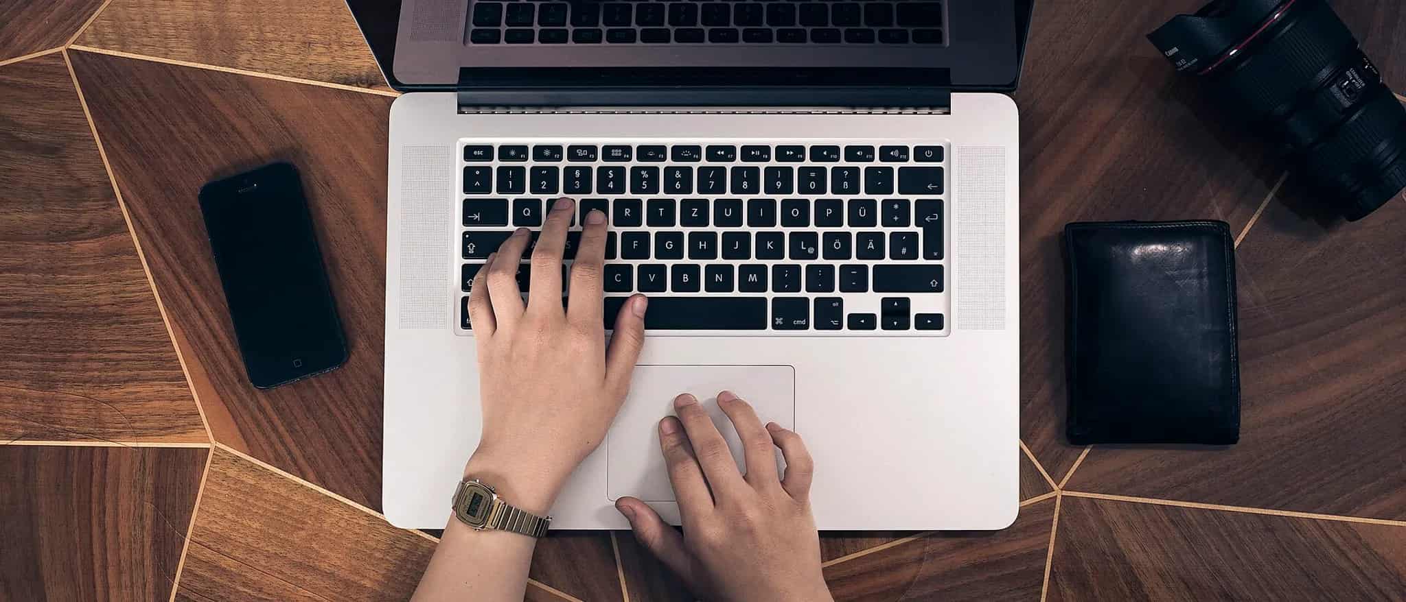 Overhead view of two hands typing on a laptop keyboard next to a smartphone on a wooden desk.