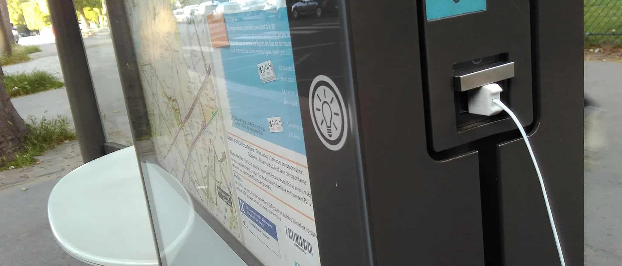 Close-up of a white USB cable plugged into a public charging port built into a city bus stop shelter.