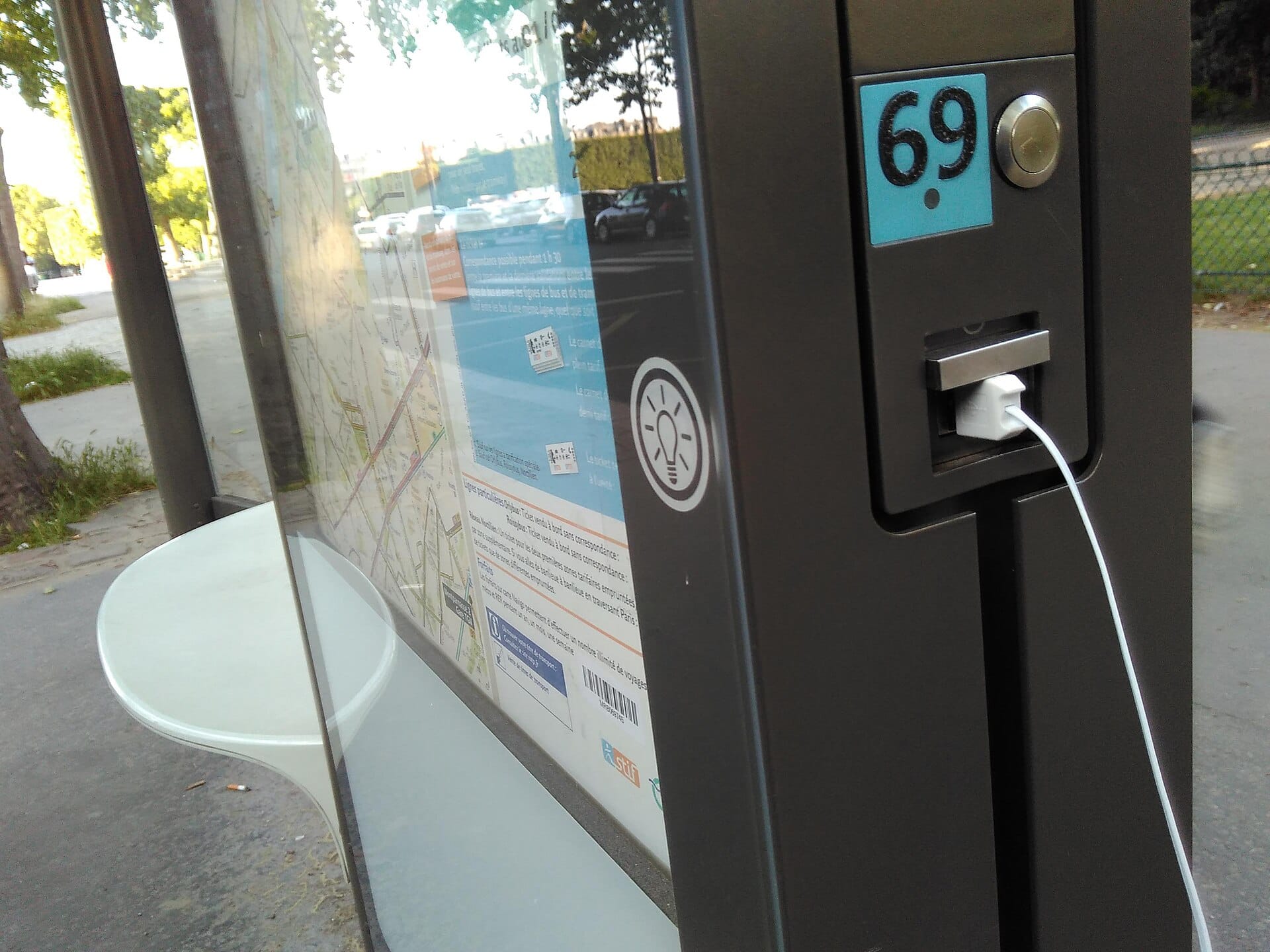 Close-up of a white USB cable plugged into a public charging port built into a city bus stop shelter.