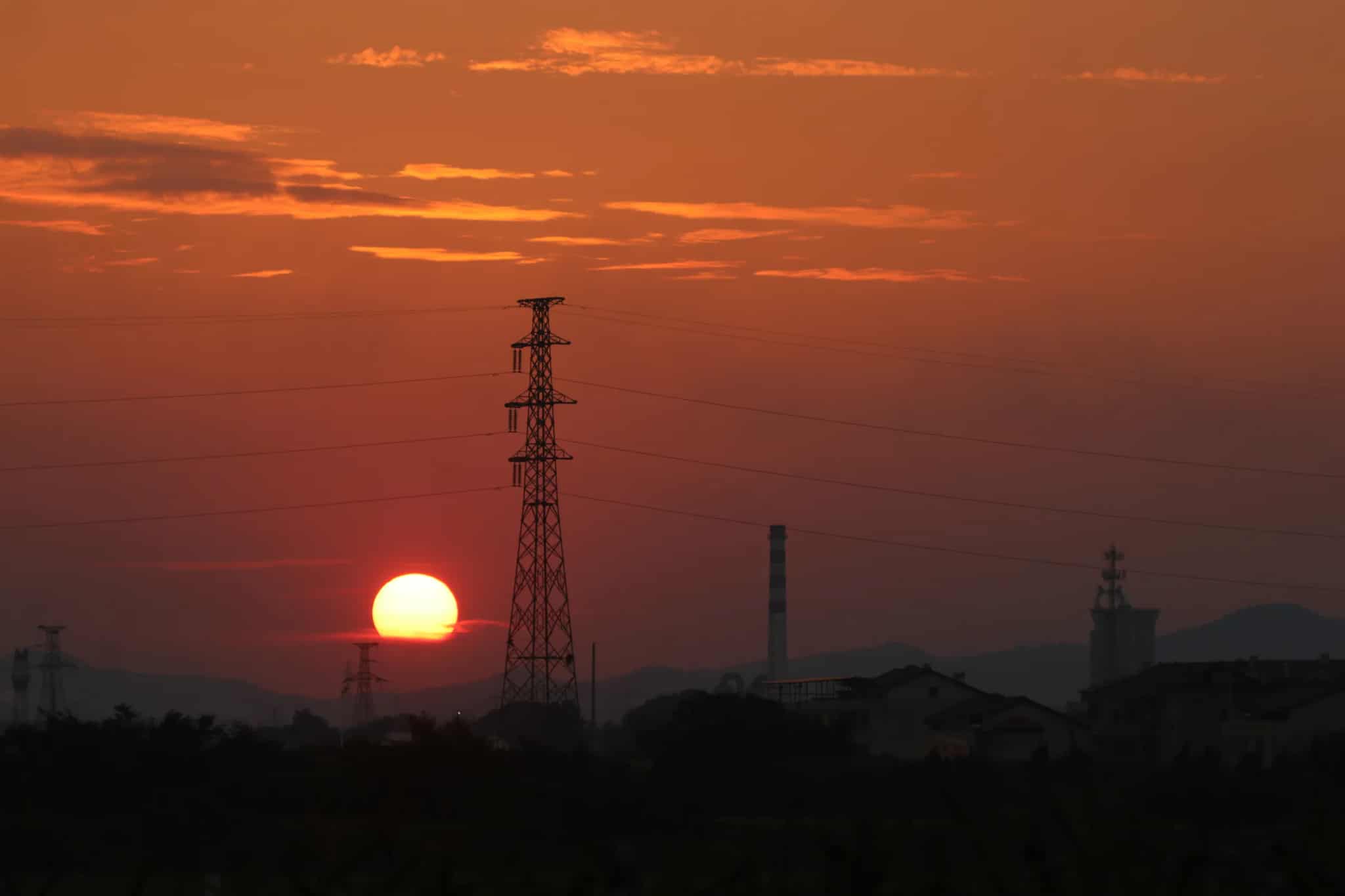 Silhouette of a high-voltage electrical transmission tower and power lines against a vivid orange sunset sky.