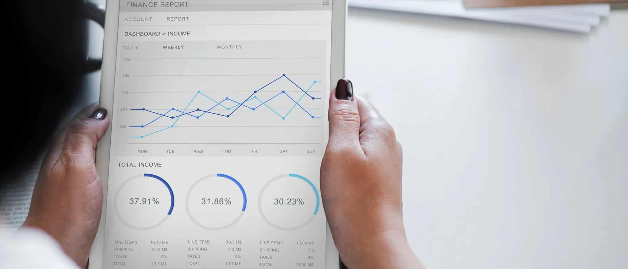 A pair of hands holds a tablet displaying a finance report dashboard with line graphs and percentage charts.