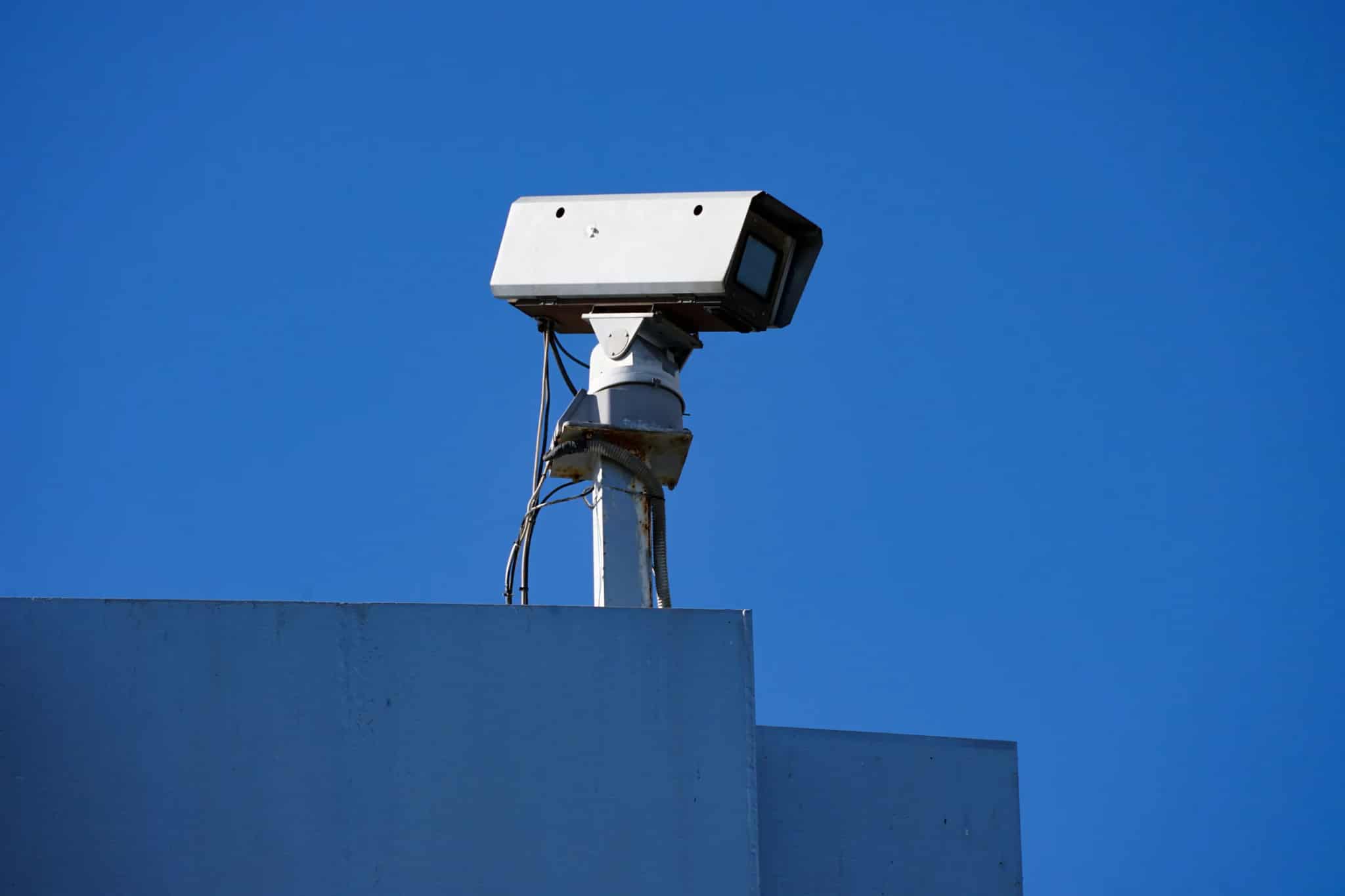 A security camera mounted on top of a building, viewed from below against a clear deep-blue sky.