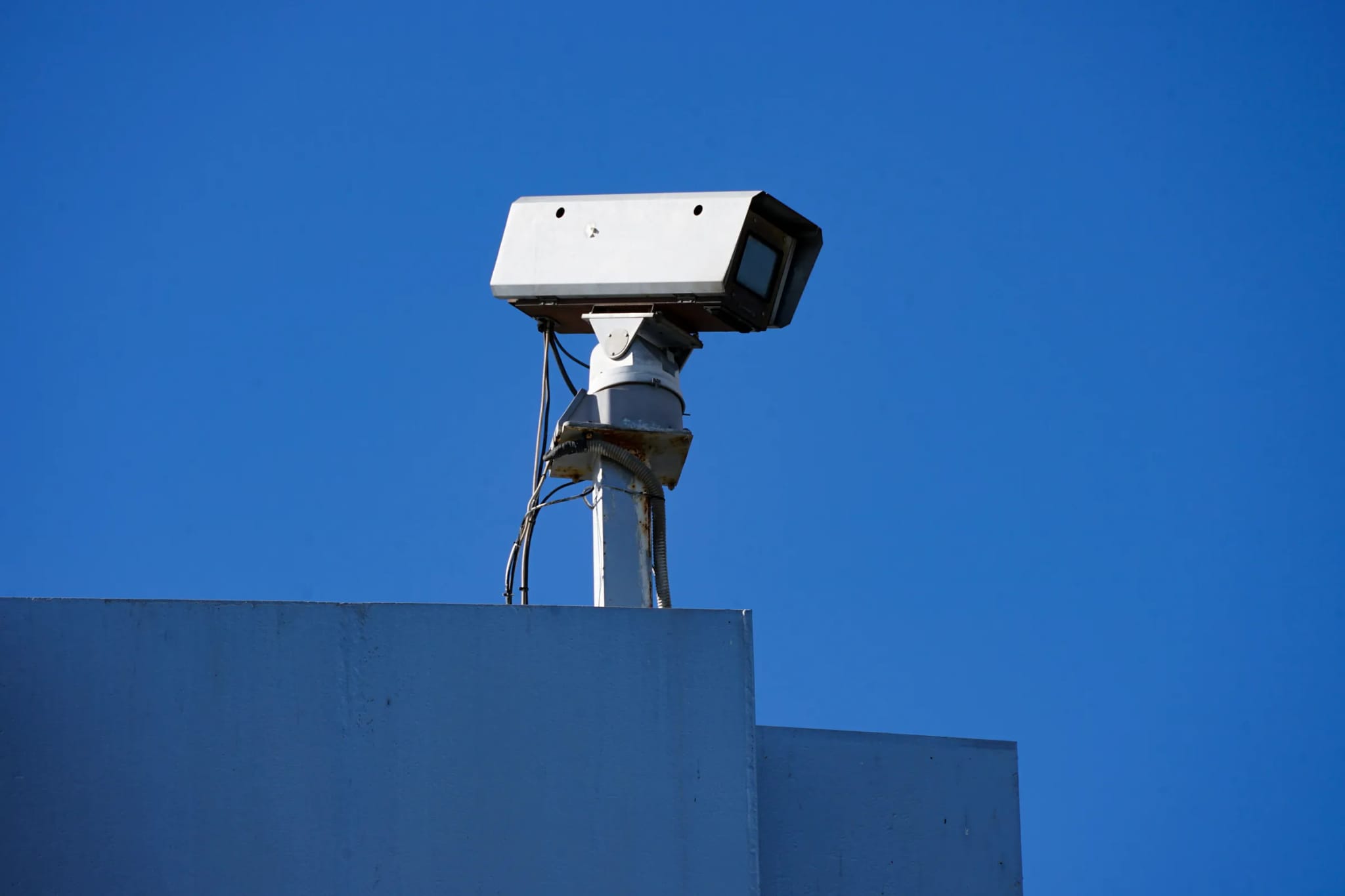 A security camera mounted on top of a building, viewed from below against a clear deep-blue sky.
