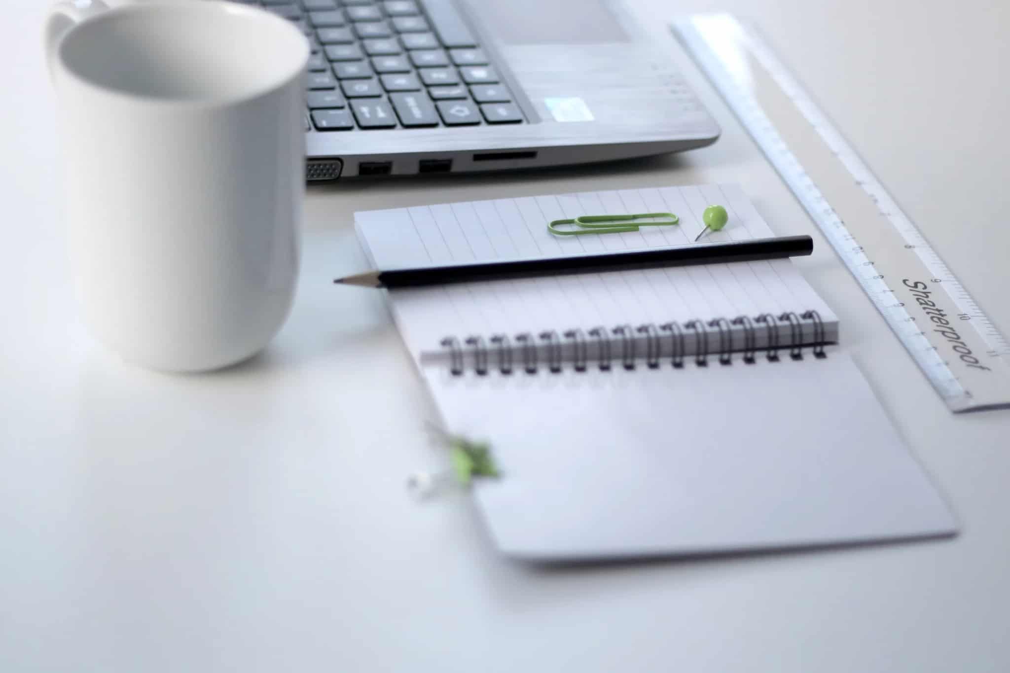 An organized desk flat-lay with a laptop, spiral notebook, pencil, coffee mug, and ruler on a white surface.