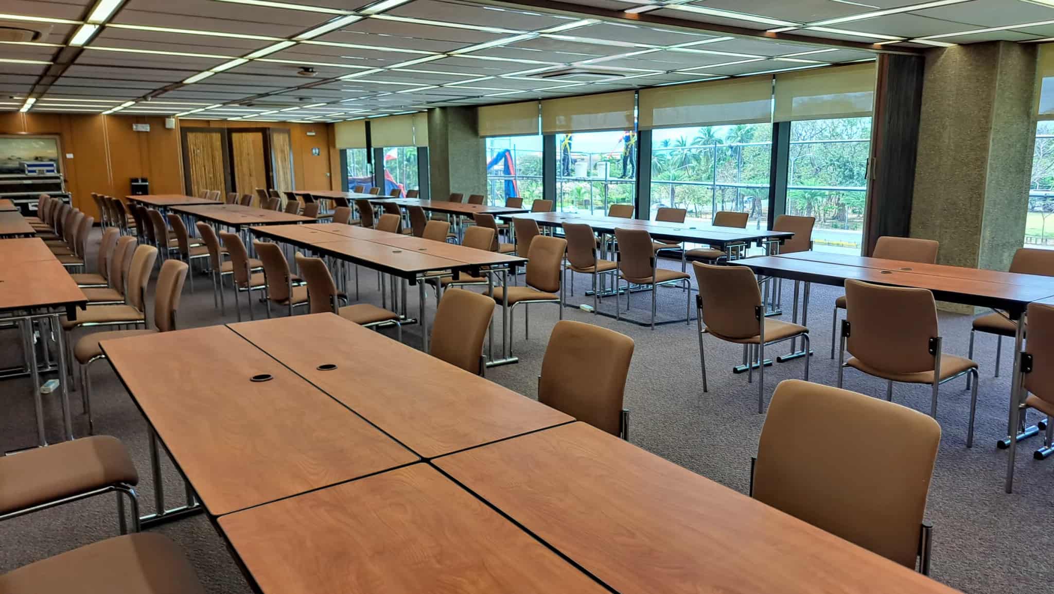 A spacious, empty conference room with long rows of wooden tables and beige chairs, lit by large windows.