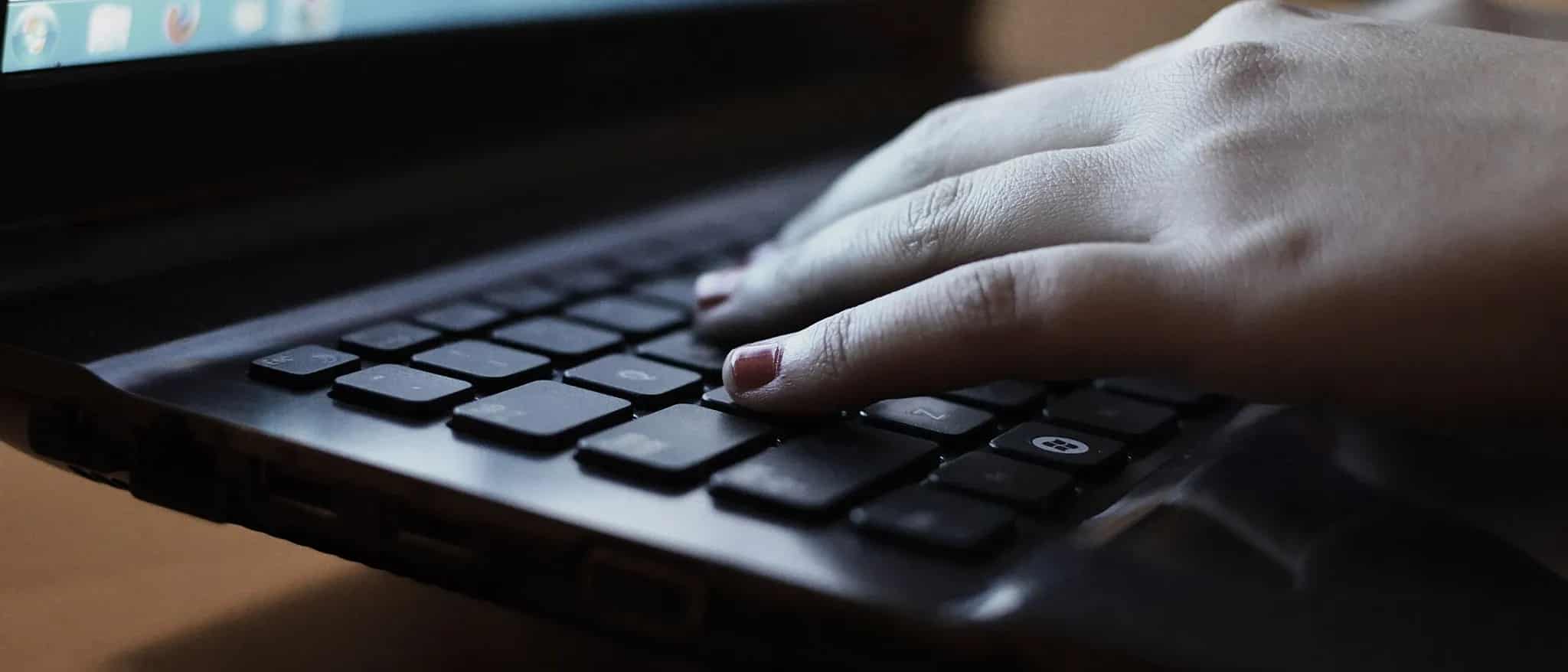 Close-up of a hand typing on a laptop keyboard with a blurred screen glowing in the background.