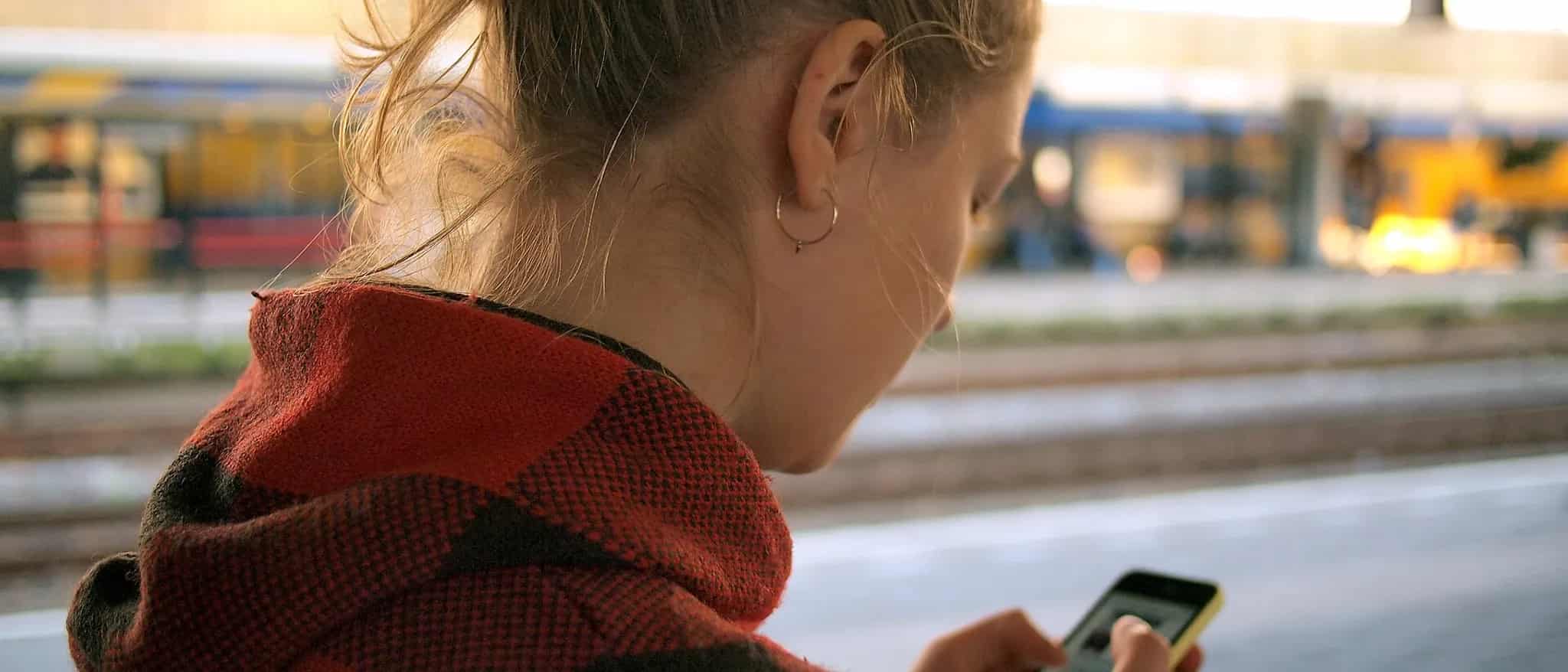 A person seen from behind checks a smartphone while waiting outdoors at a train platform in warm evening light.