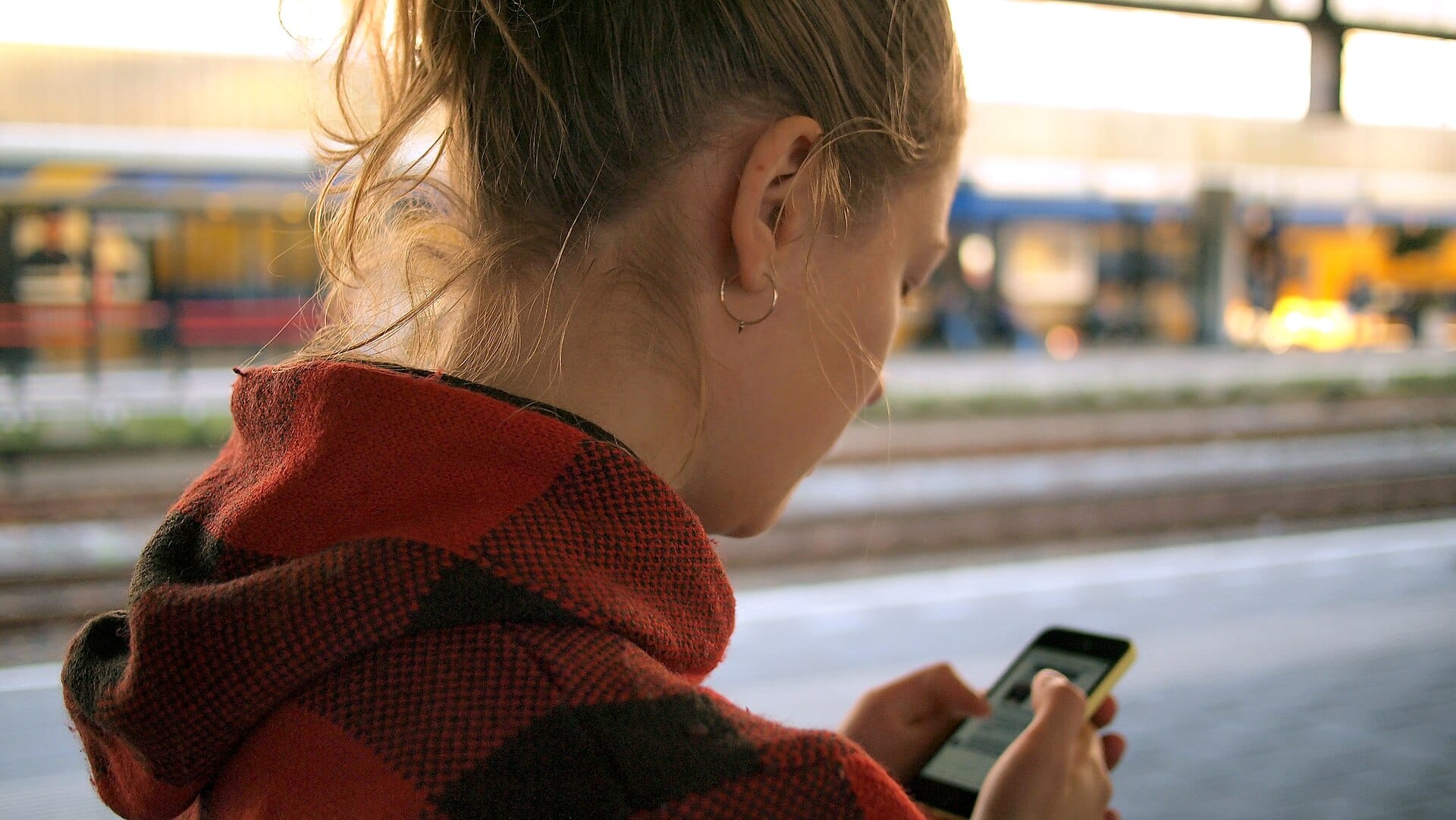 A person seen from behind checks a smartphone while waiting outdoors at a train platform in warm evening light.