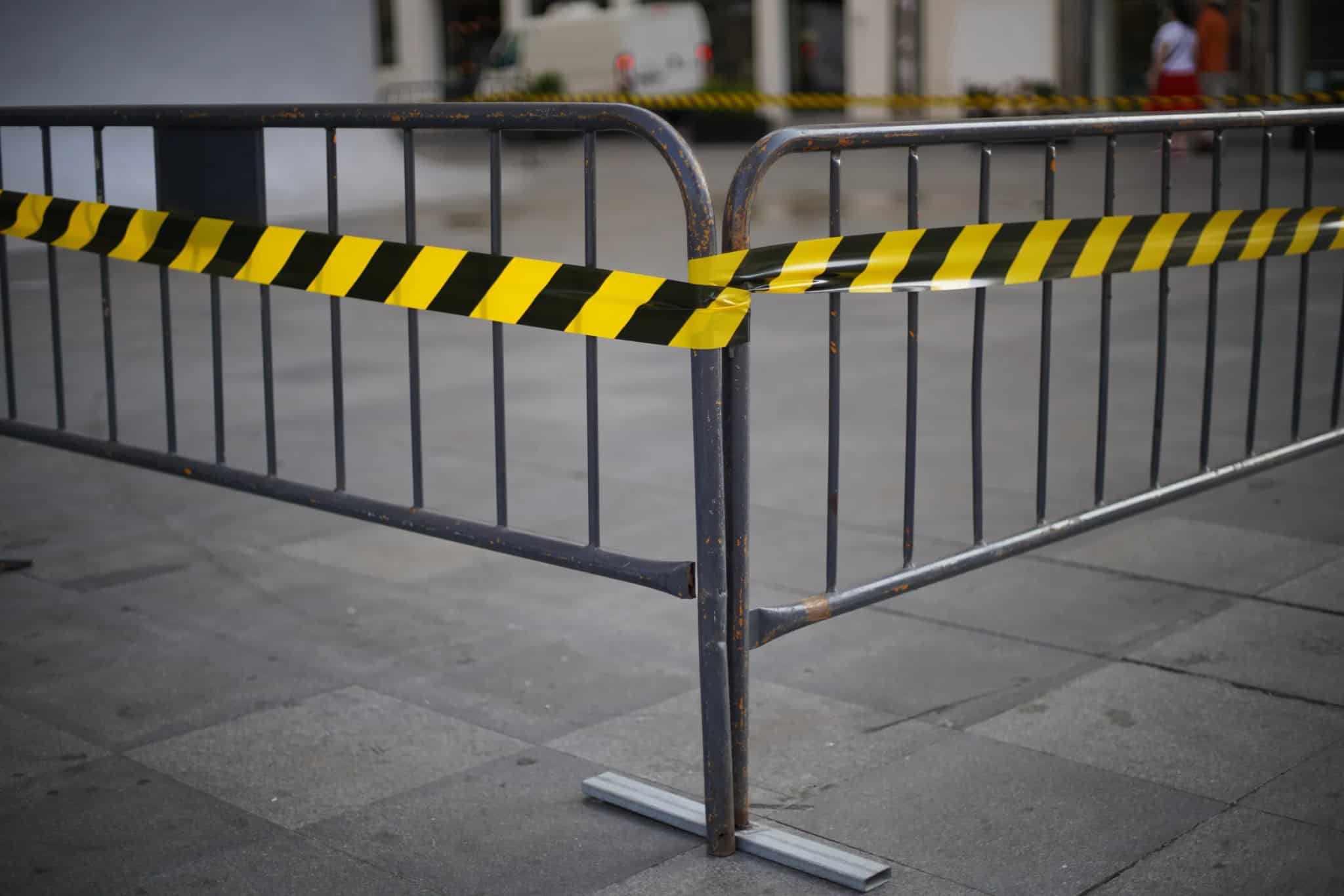 A metal crowd barricade wrapped in black-and-yellow caution tape on a paved plaza.