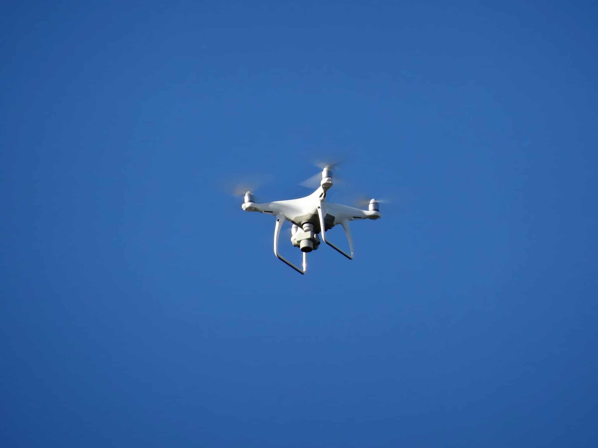 A white quadcopter drone hovering mid-flight against a clear blue sky.