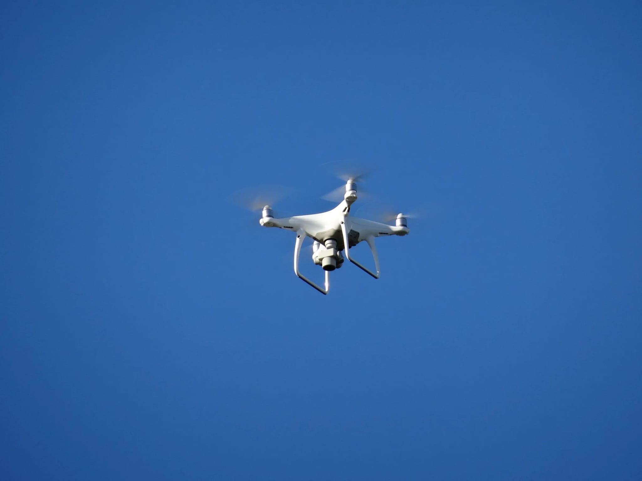 A white quadcopter drone hovering mid-flight against a clear blue sky.