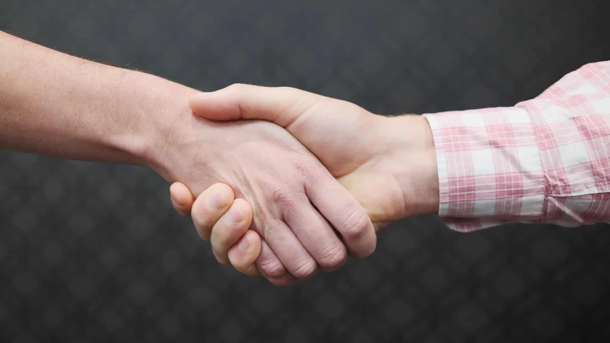 Close-up of two people shaking hands, forearms only, against a dark background.