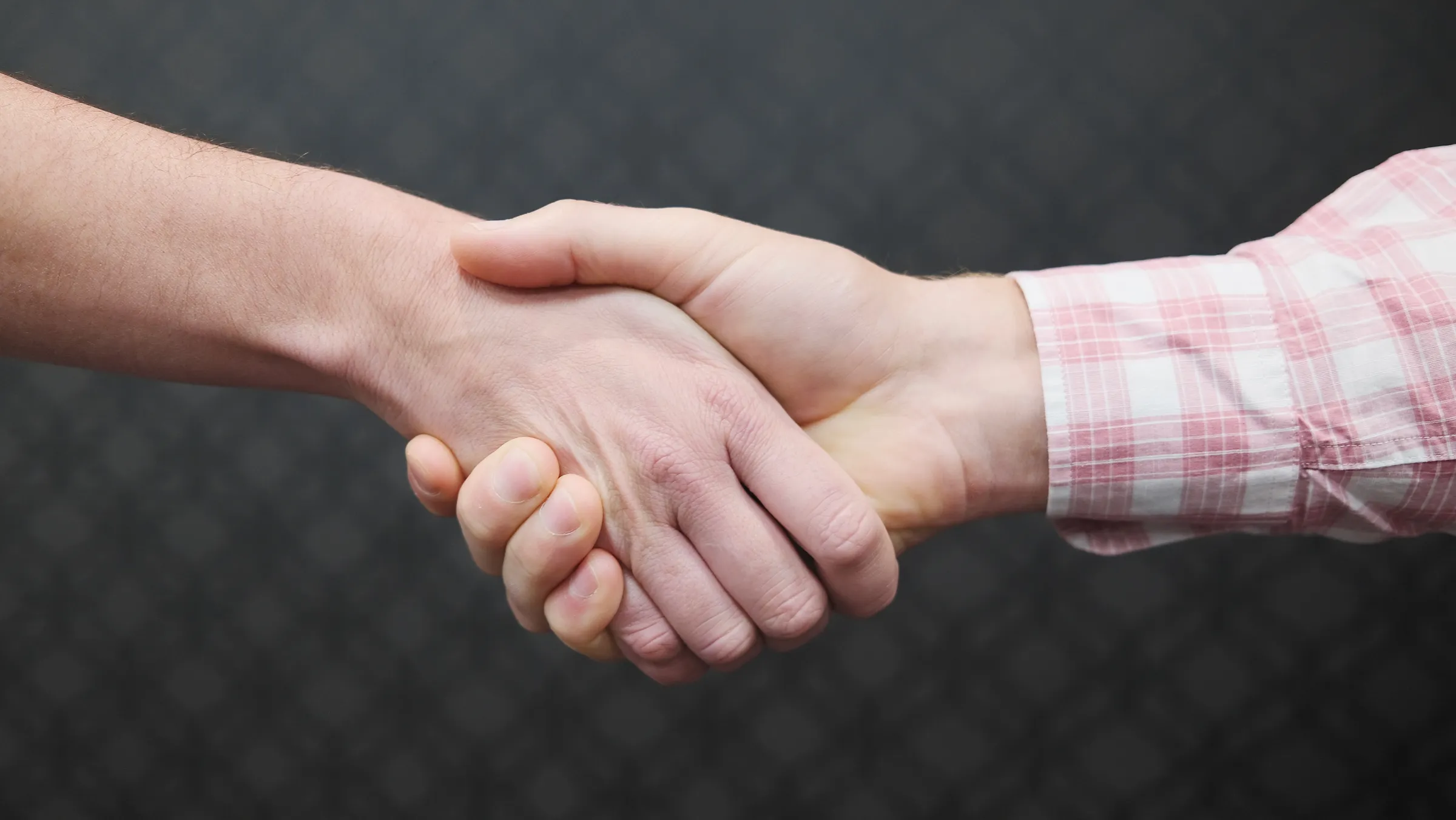 Close-up of two people shaking hands, forearms only, against a dark background.