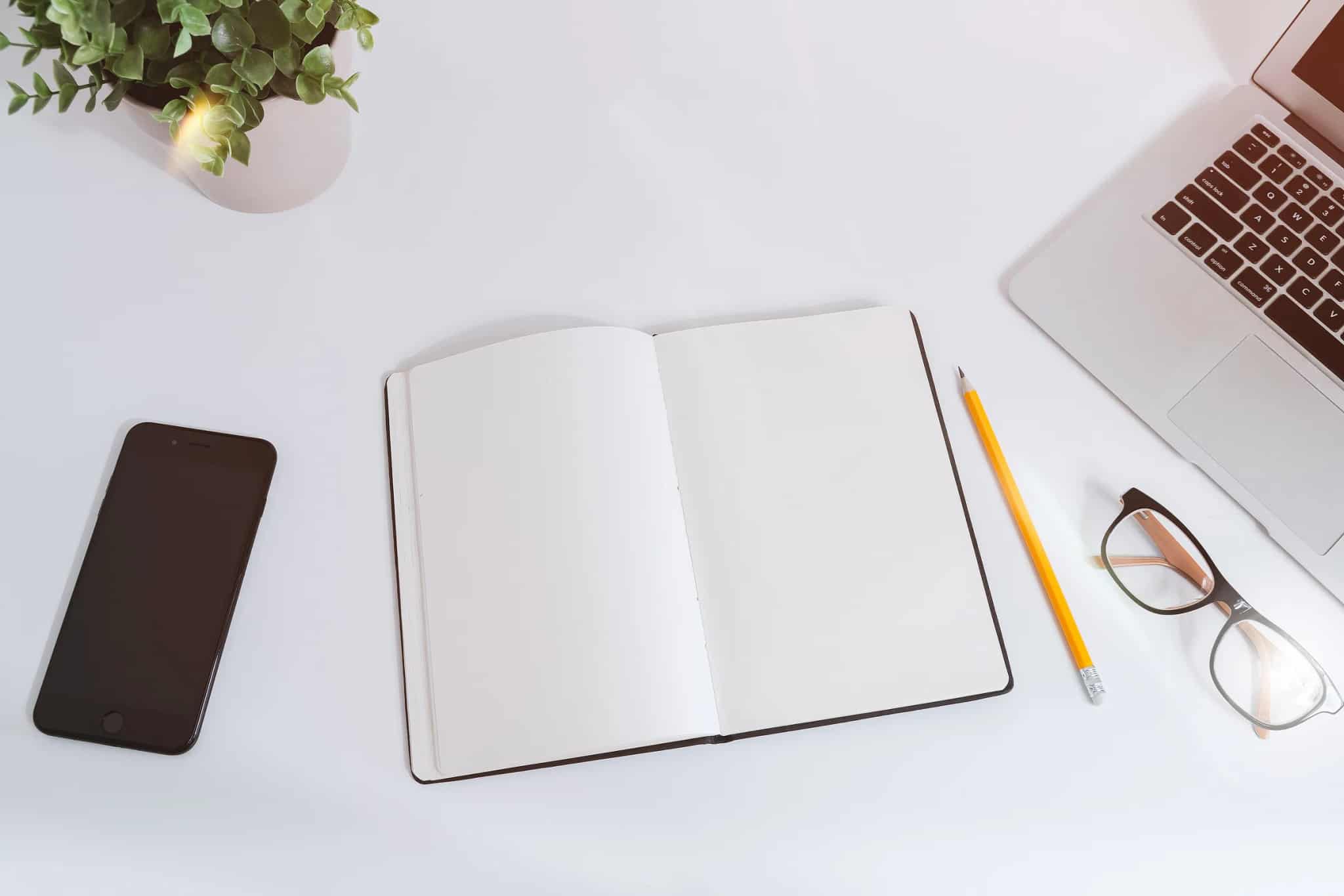 A bright overhead flat-lay of a smartphone, an open blank notebook, a pencil, glasses, a small plant, and a laptop edge on a white desk.