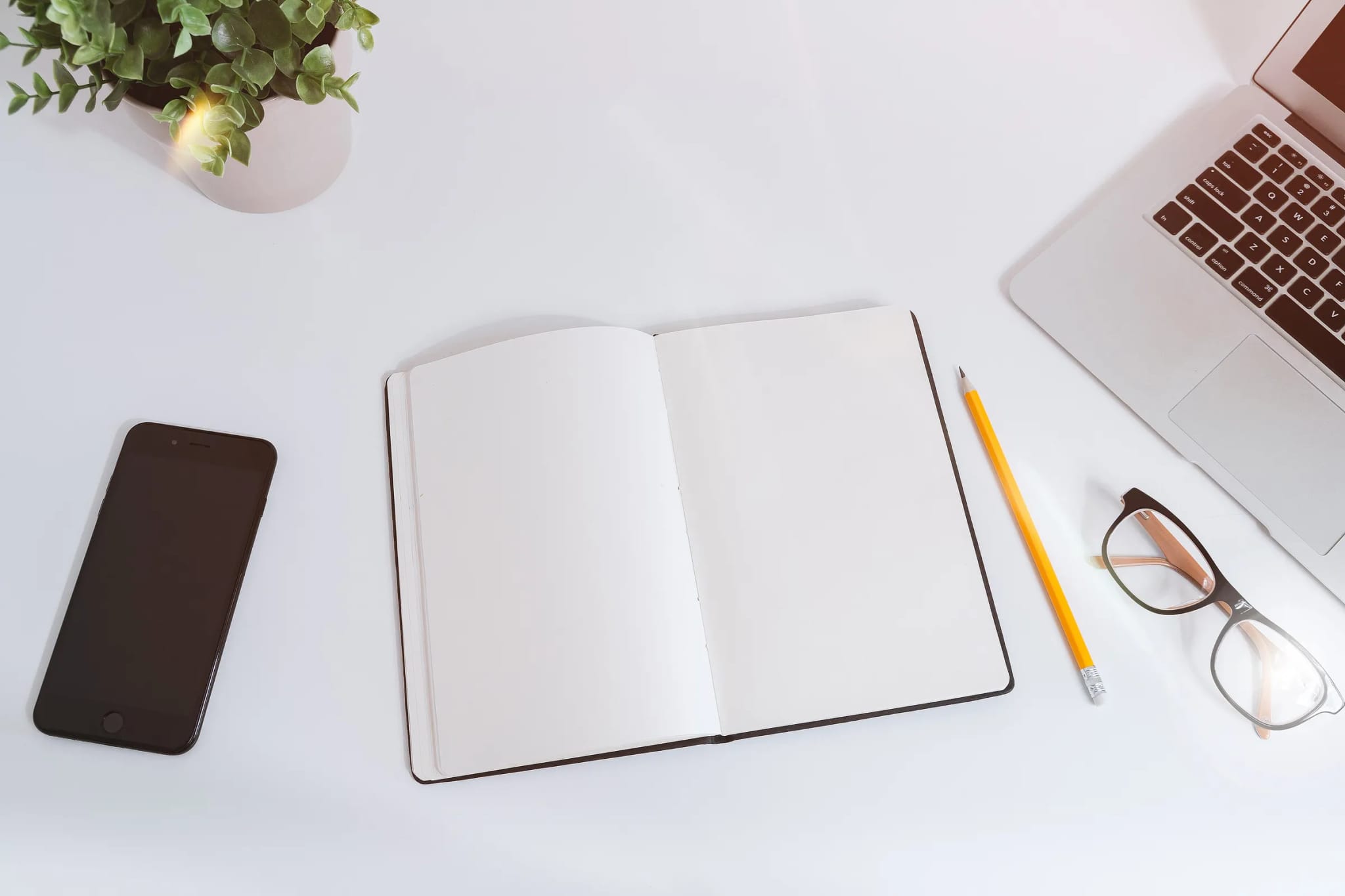A bright overhead flat-lay of a smartphone, an open blank notebook, a pencil, glasses, a small plant, and a laptop edge on a white desk.