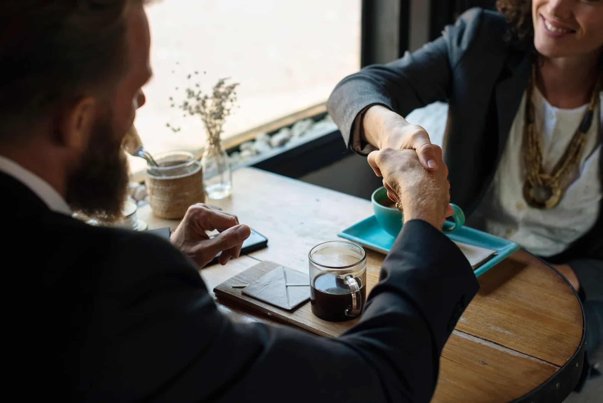 Two professionals shake hands across a wooden cafe table with coffee cups and a small vase of flowers between them.