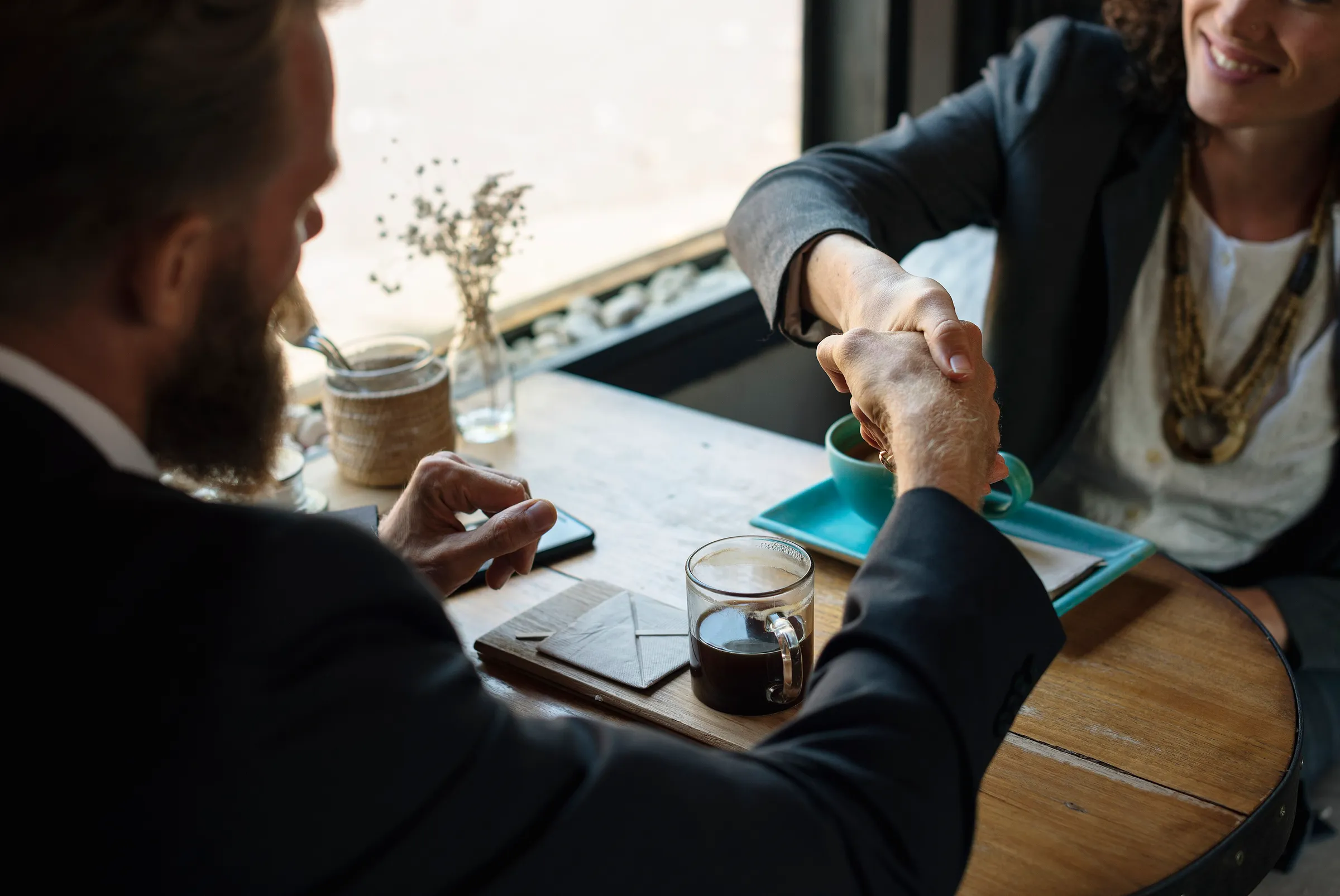 Two professionals shake hands across a wooden cafe table with coffee cups and a small vase of flowers between them.