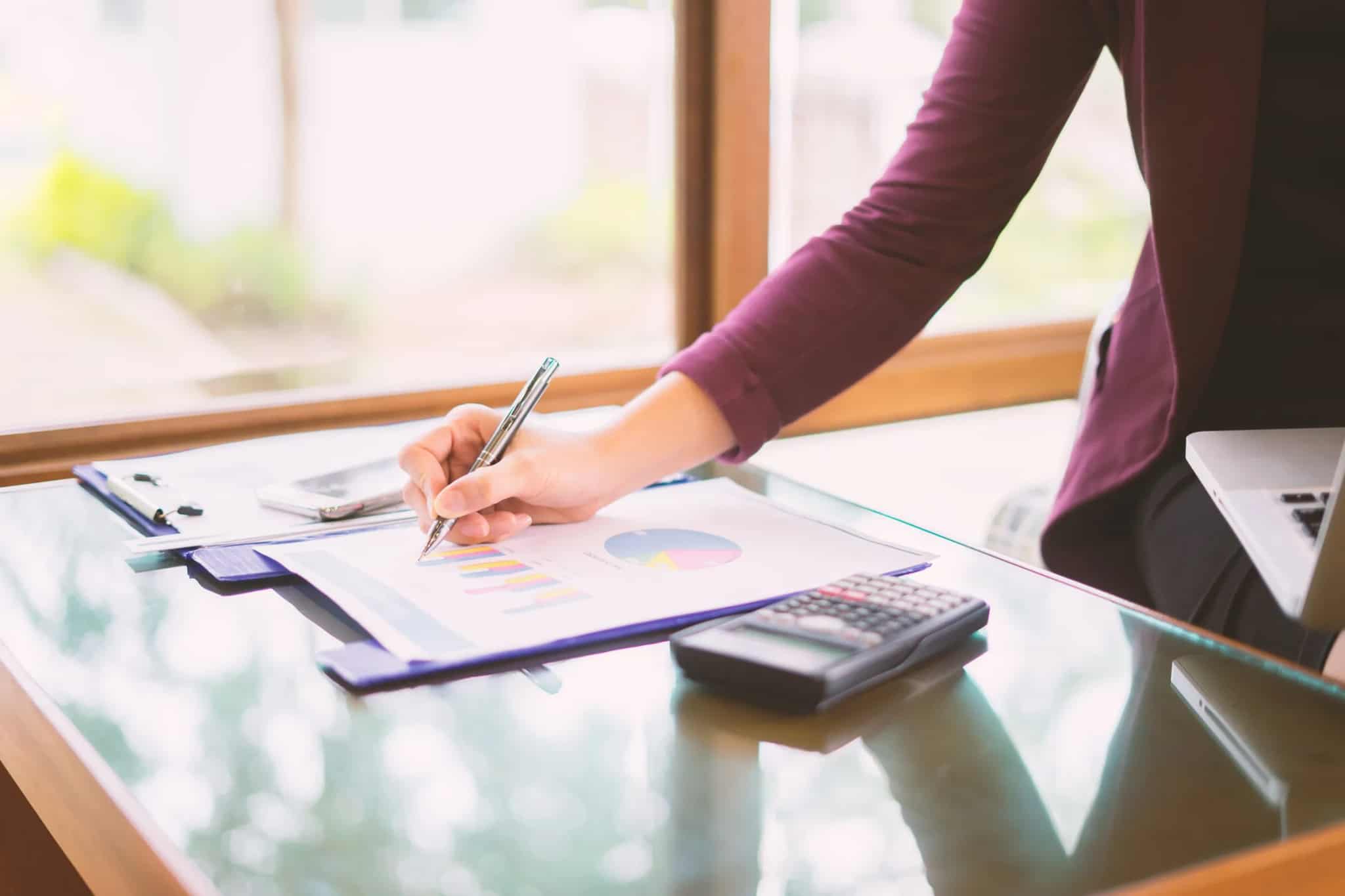 A hand holding a pen marks up a printed bar and pie chart report beside a calculator on a glass desk.