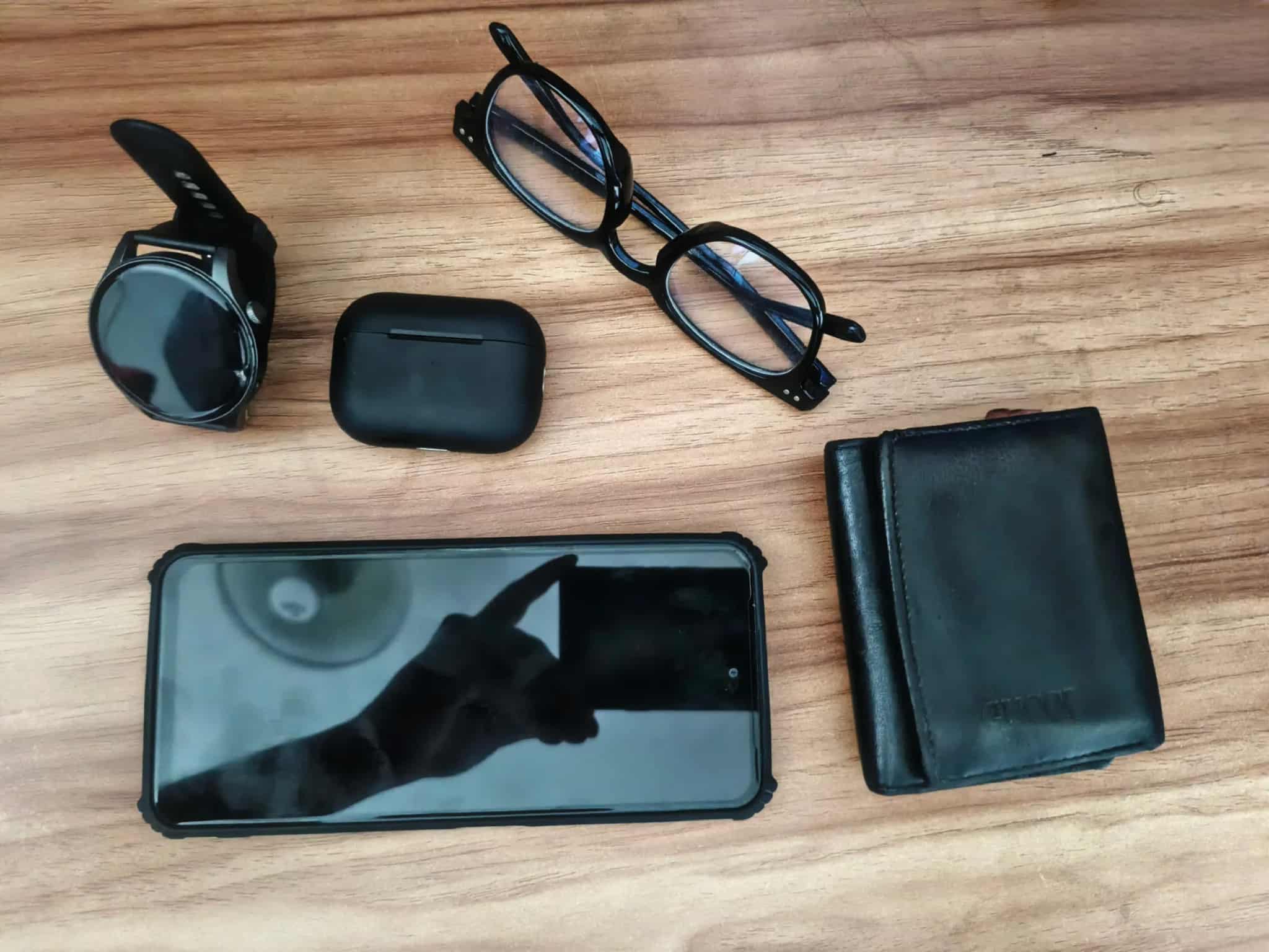 A smartphone, smartwatch, wireless earbud case, eyeglasses and wallet arranged on a wood surface.