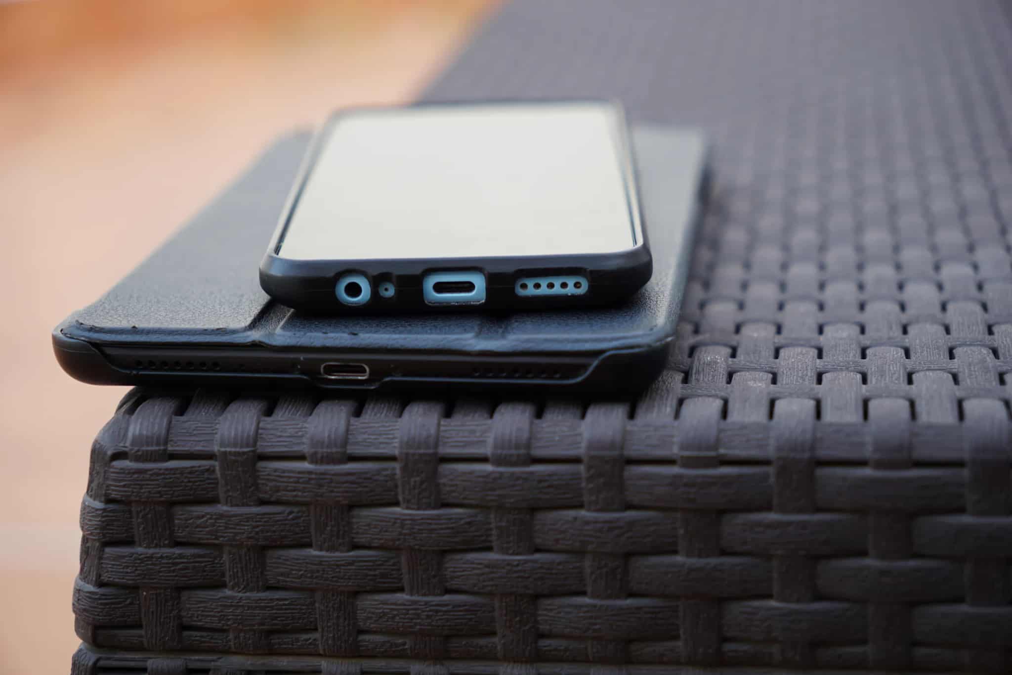 A close-up macro view of a smartphone's charging port and headphone jack resting against a tablet on a dark woven table.