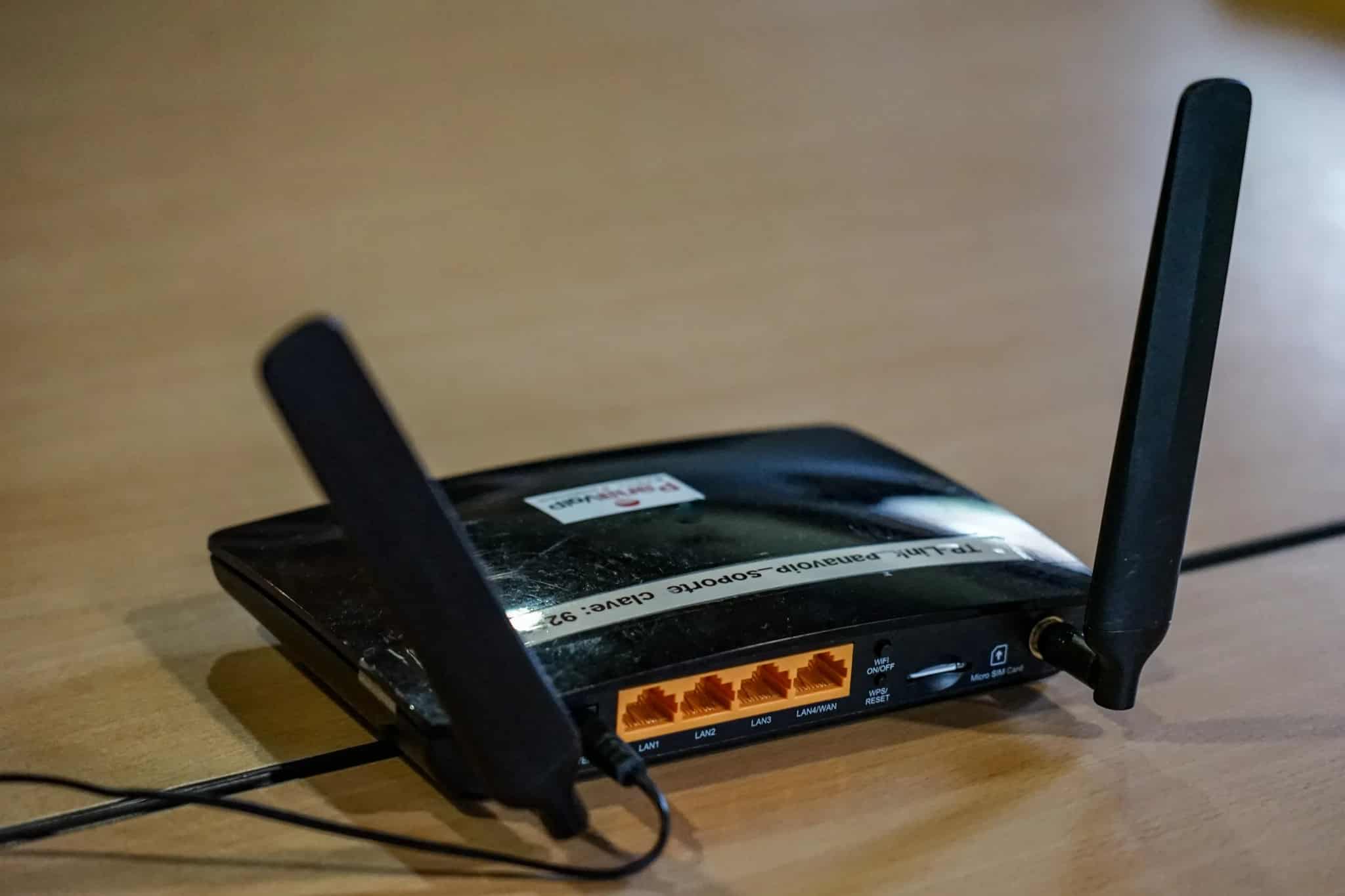 A black wireless router with two upright antennas sits on a wooden table with a cable plugged into its back panel.
