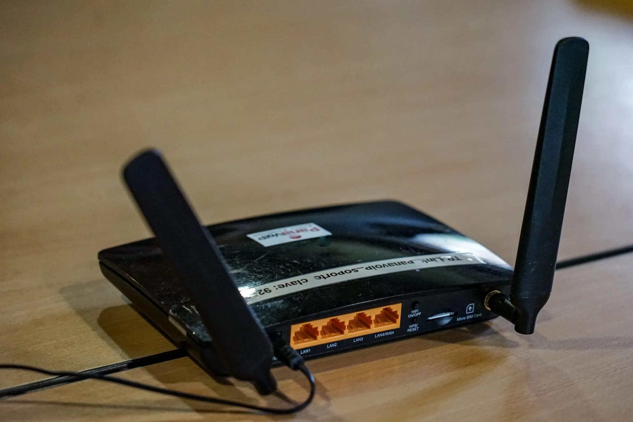 A black wireless router with two upright antennas sits on a wooden table with a cable plugged into its back panel.