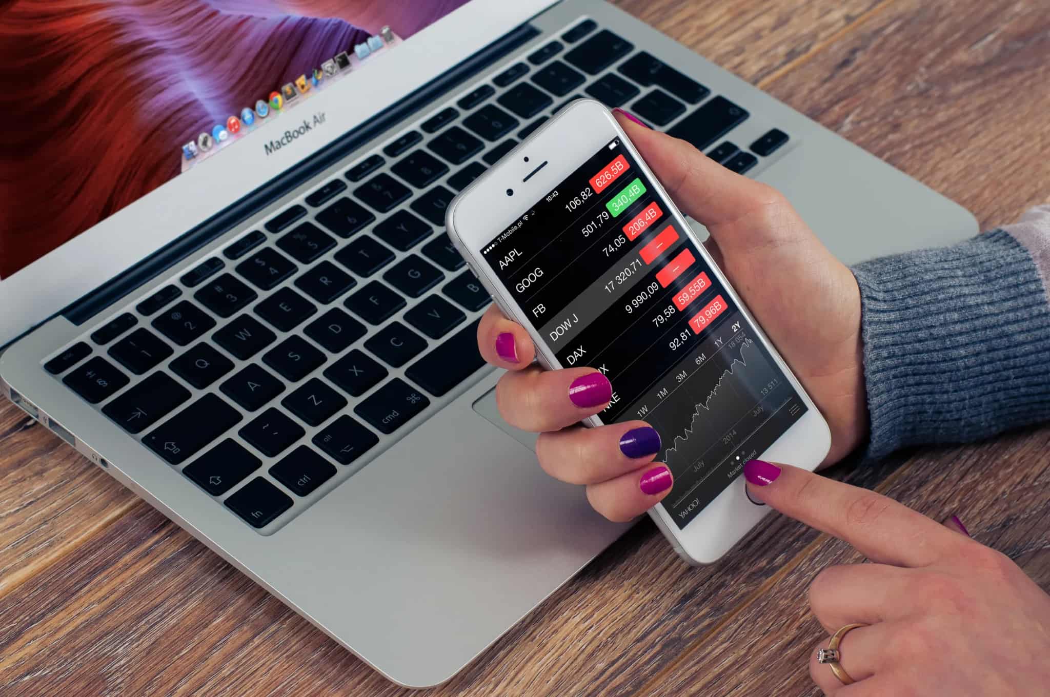 A hand holds a smartphone displaying a financial market-tracking app with stock tickers and a price chart, beside an open laptop keyboard.