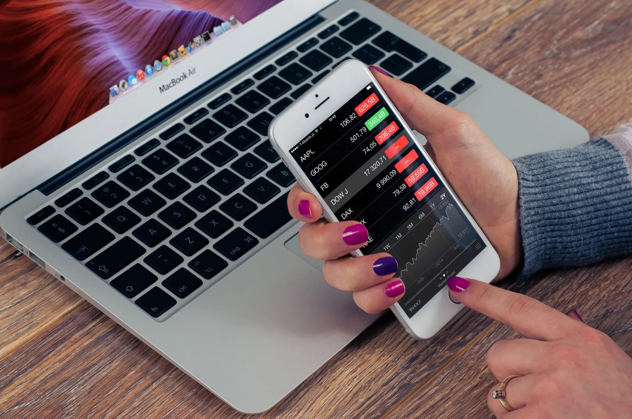 A hand holds a smartphone displaying a financial market-tracking app with stock tickers and a price chart, beside an open laptop keyboard.