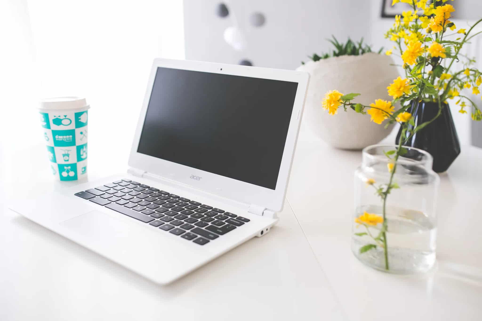 A white laptop with its screen off sitting on a desk beside a coffee cup and a vase of yellow flowers.