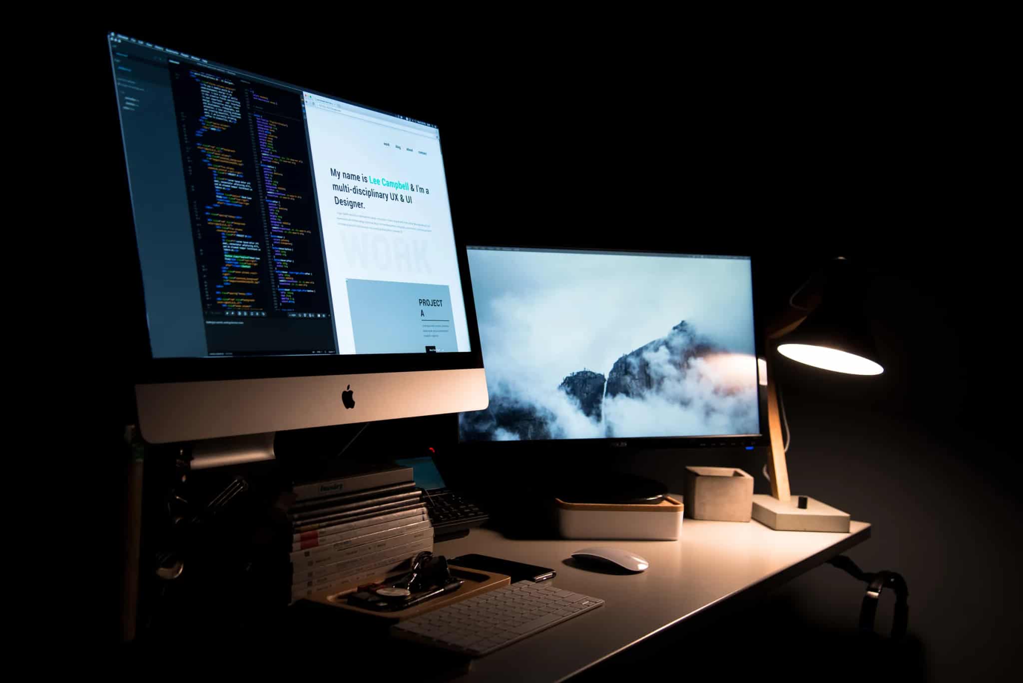 A dark home office at night with a two-monitor setup: one screen shows a code editor and a personal portfolio website, the other a misty mountain photo, lit by a warm desk lamp.
