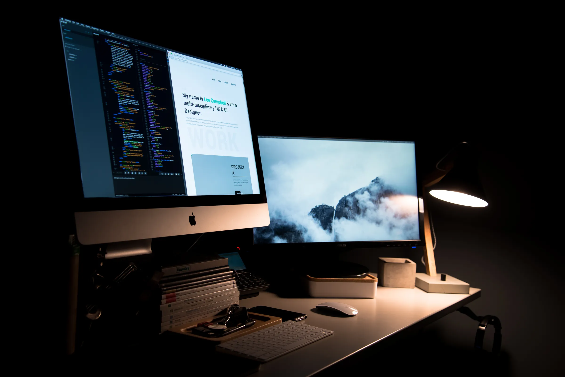 A dark home office at night with a two-monitor setup: one screen shows a code editor and a personal portfolio website, the other a misty mountain photo, lit by a warm desk lamp.