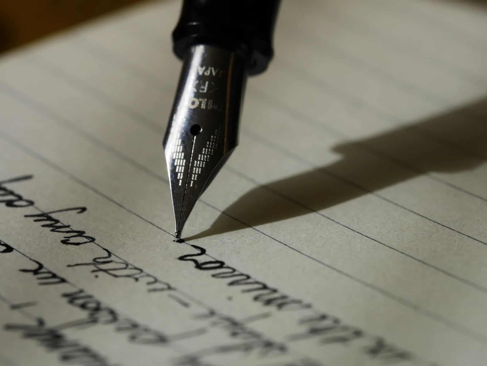 Close-up of a fountain pen nib writing cursive script on lined paper, its shadow falling across the page.